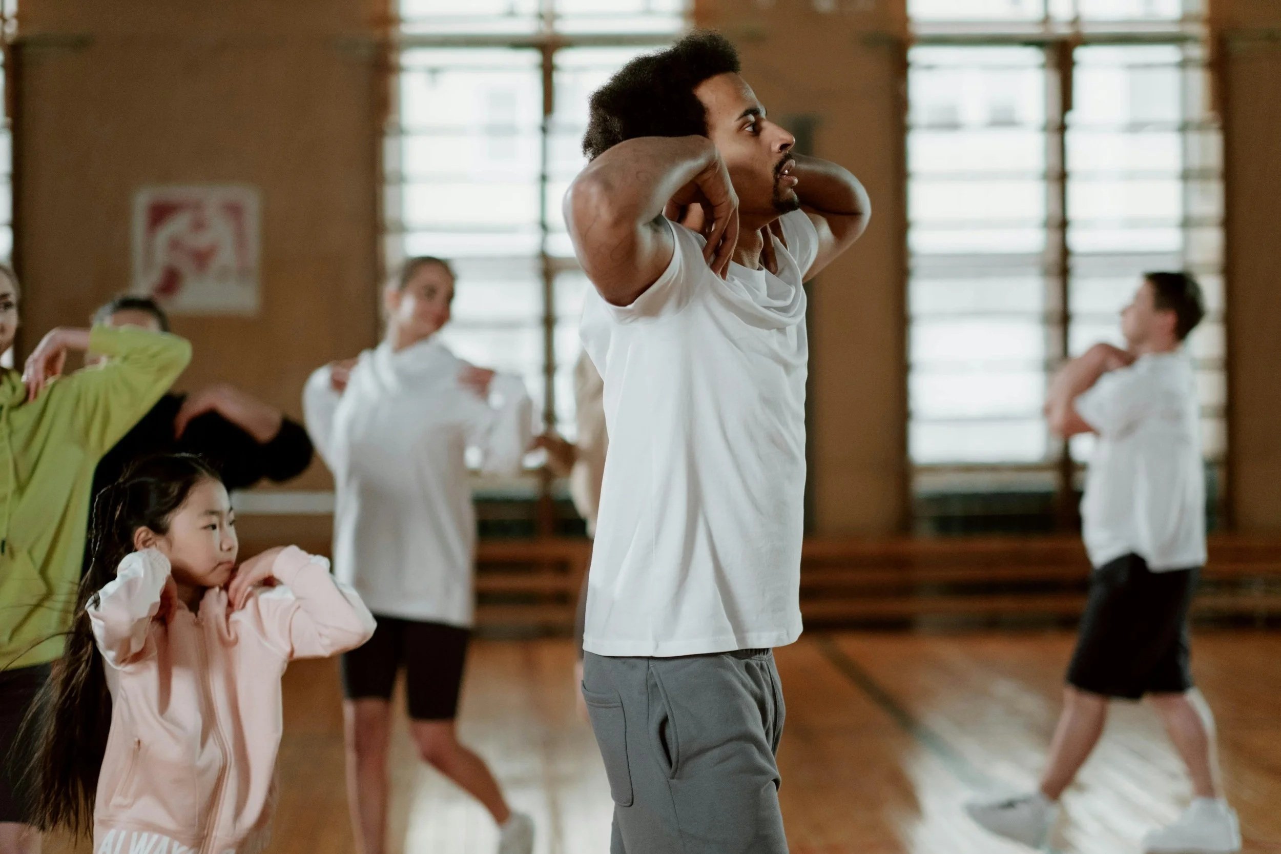 A man standing in a gym or dance studio with his hands behind his head, practicing stretching or warm-up exercises, surrounded by a group of children and women also in workout poses.
