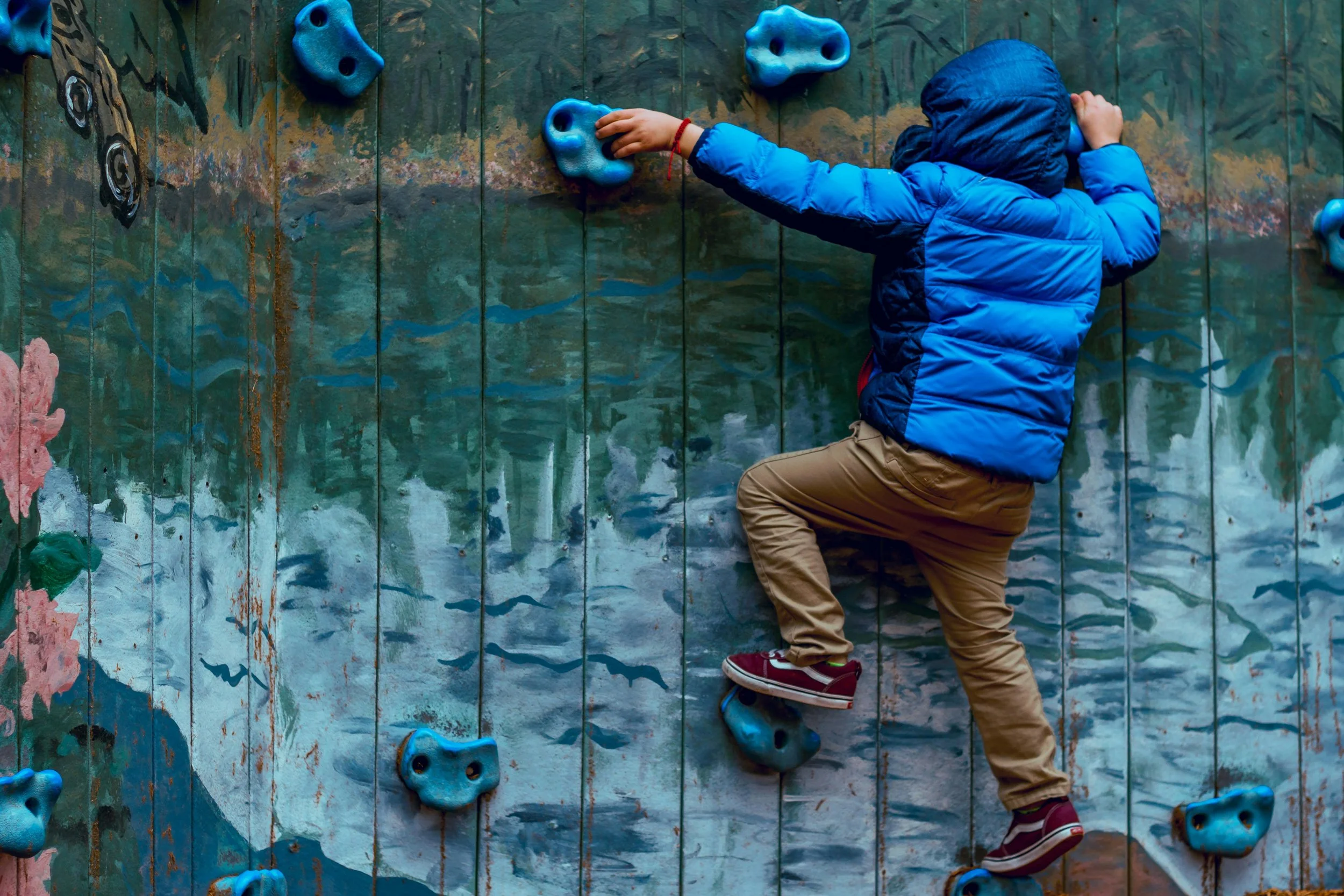 Child in blue jacket and tan pants climbing an indoor rock wall with blue holds, painted in mountain and lake scenery.