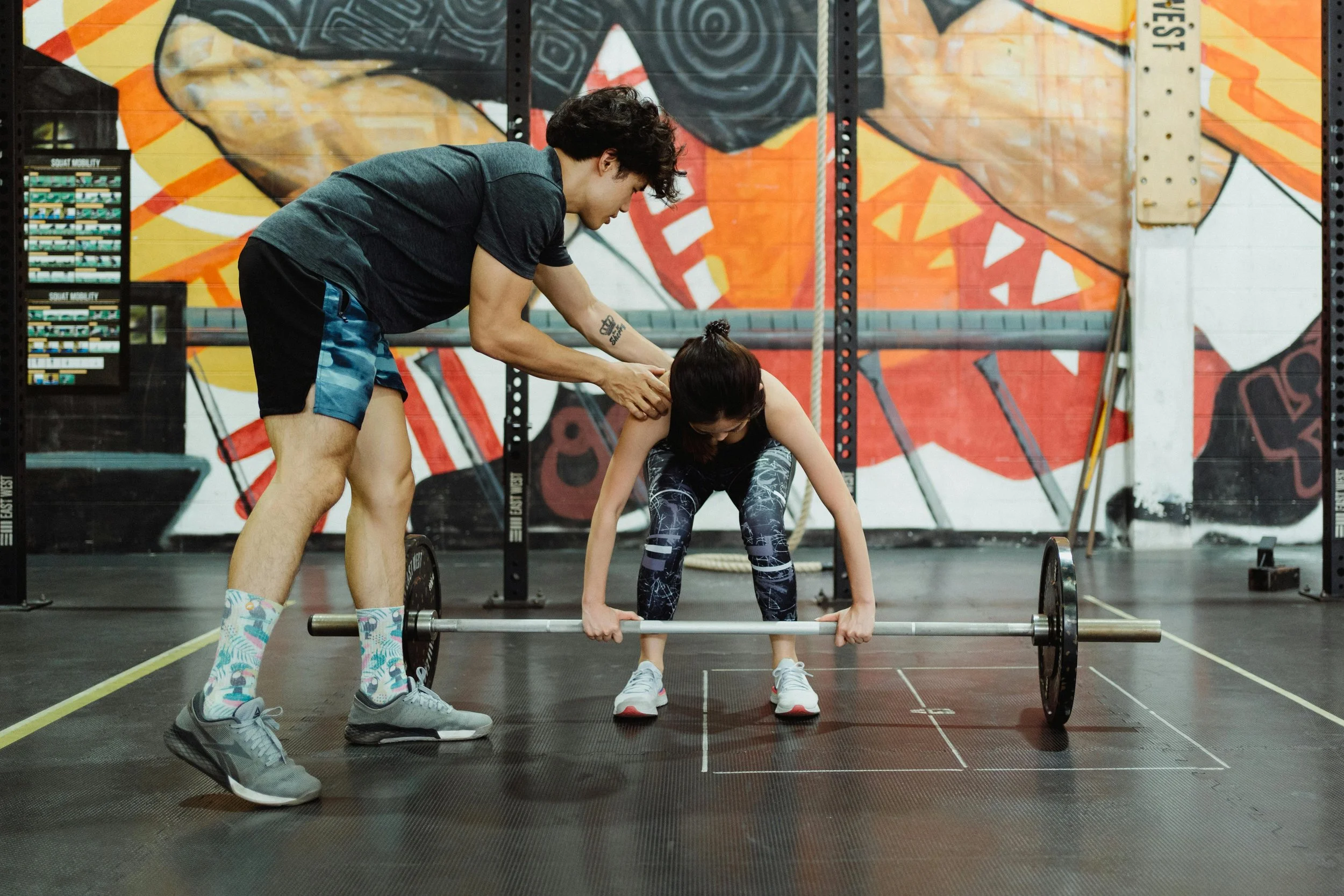 A woman lifting a barbell with the help of a man in a gym with colorful wall art and climbing ropes.