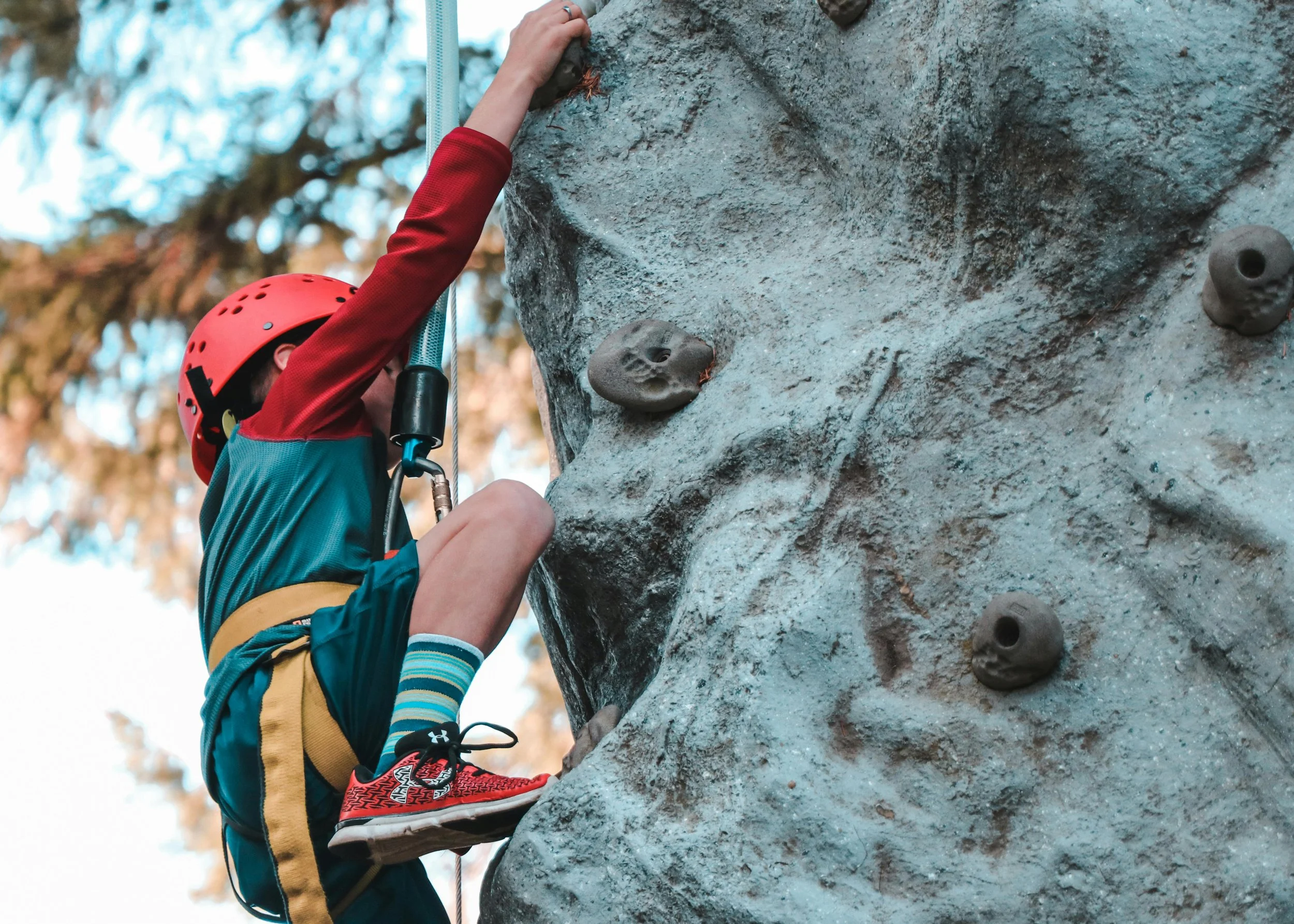Child wearing a red helmet and blue shorts climbing a gray rock wall with indoor climbing holds outdoors.