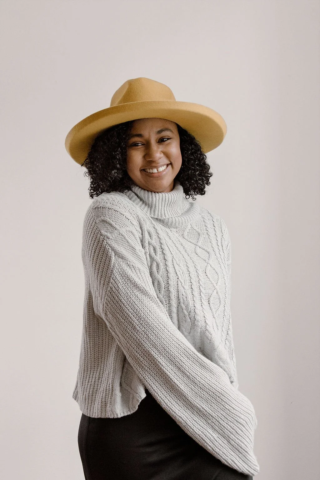 A woman smiling, wearing a beige wide-brimmed hat and a light gray cable-knit turtleneck sweater against a plain white background.