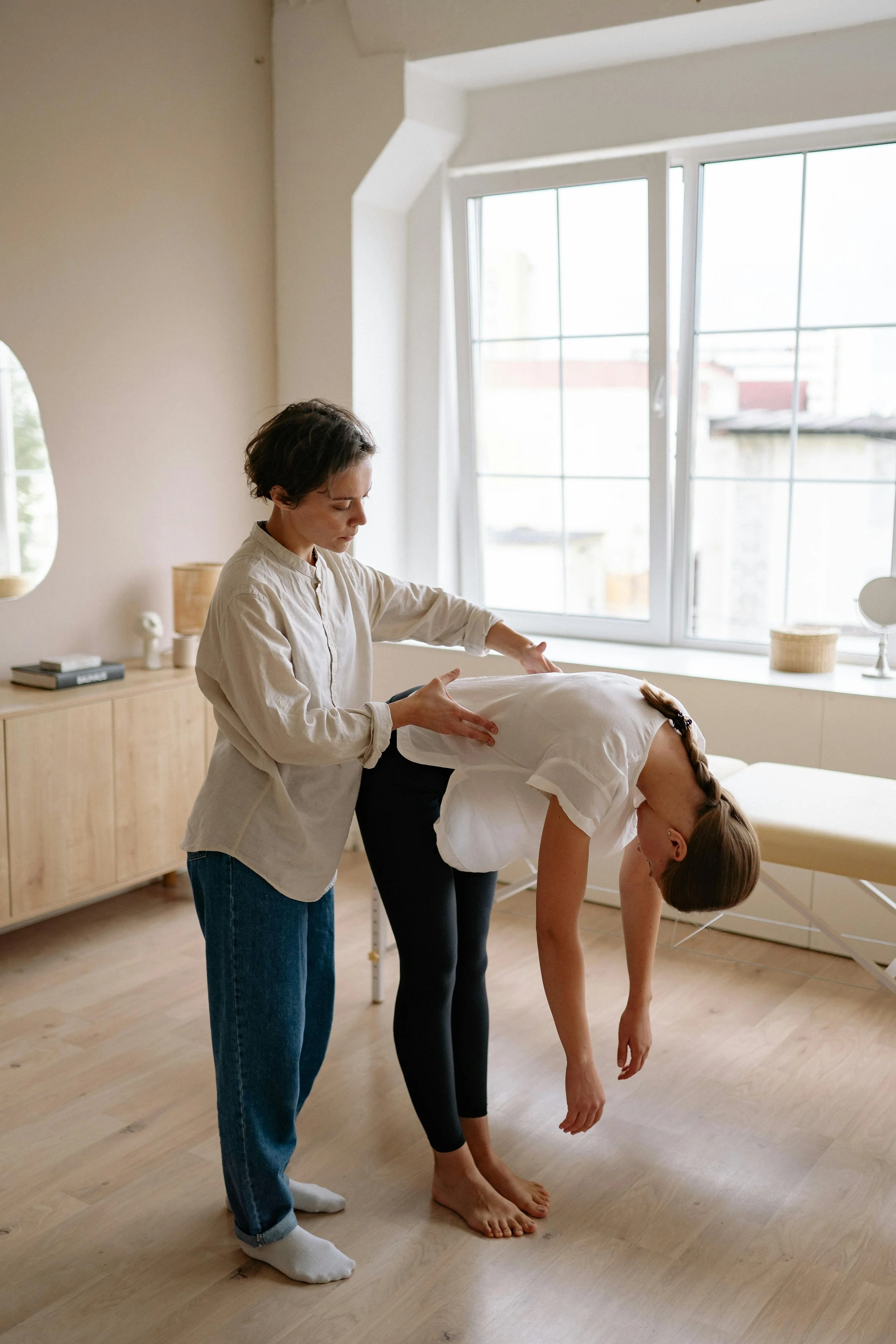 A woman is receiving a chiropractic adjustment or physical therapy from a healthcare professional in a bright, minimalistic room with wooden furniture and large windows.