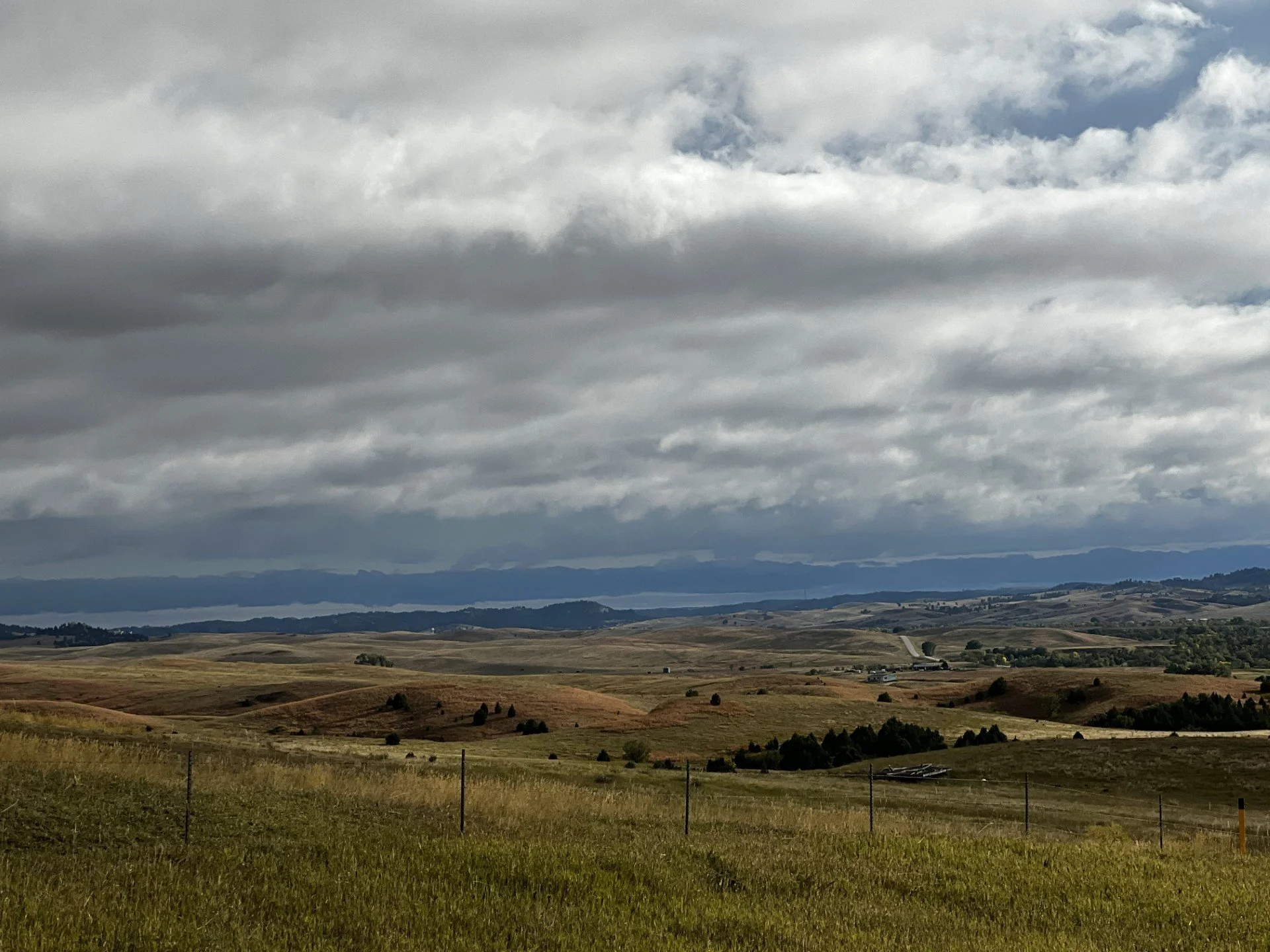 A wide view of rolling hills under a cloudy sky, with a few scattered trees and a fenced pasture in the foreground.