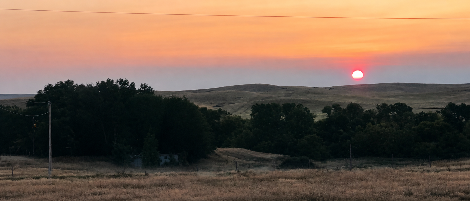 A landscape scene of rolling hills with a setting sun and colorful sky, trees in the foreground, and power lines crossing the sky.