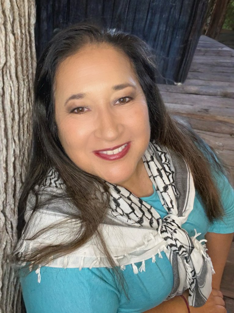 A woman smiling and leaning against a tree, with a wooden deck in the background.
