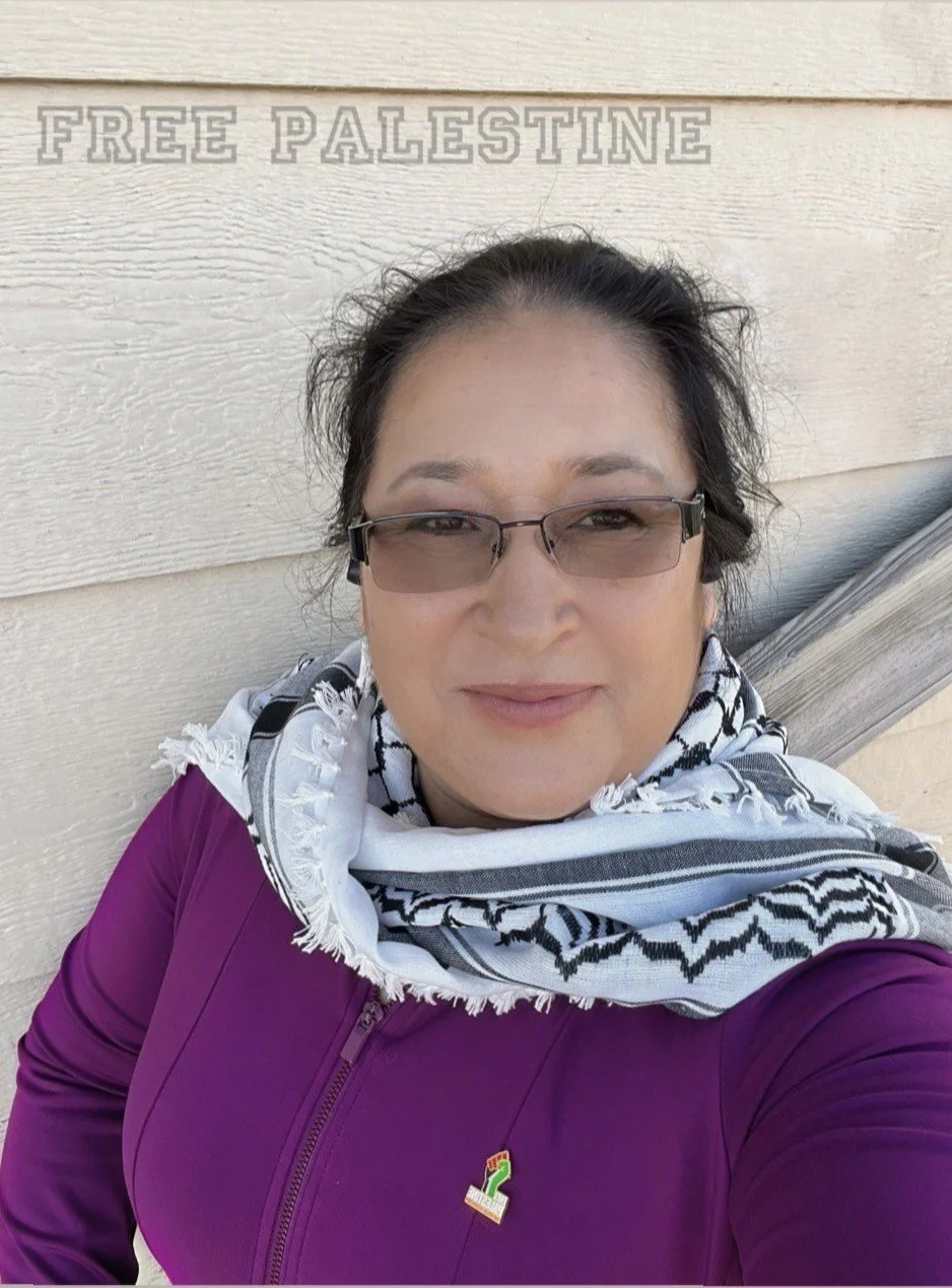 A woman with dark hair, wearing glasses, a purple jacket, and a black and white keffiyeh scarf, standing in front of a beige wall with the words 'FREE PALESTINE' in the background.