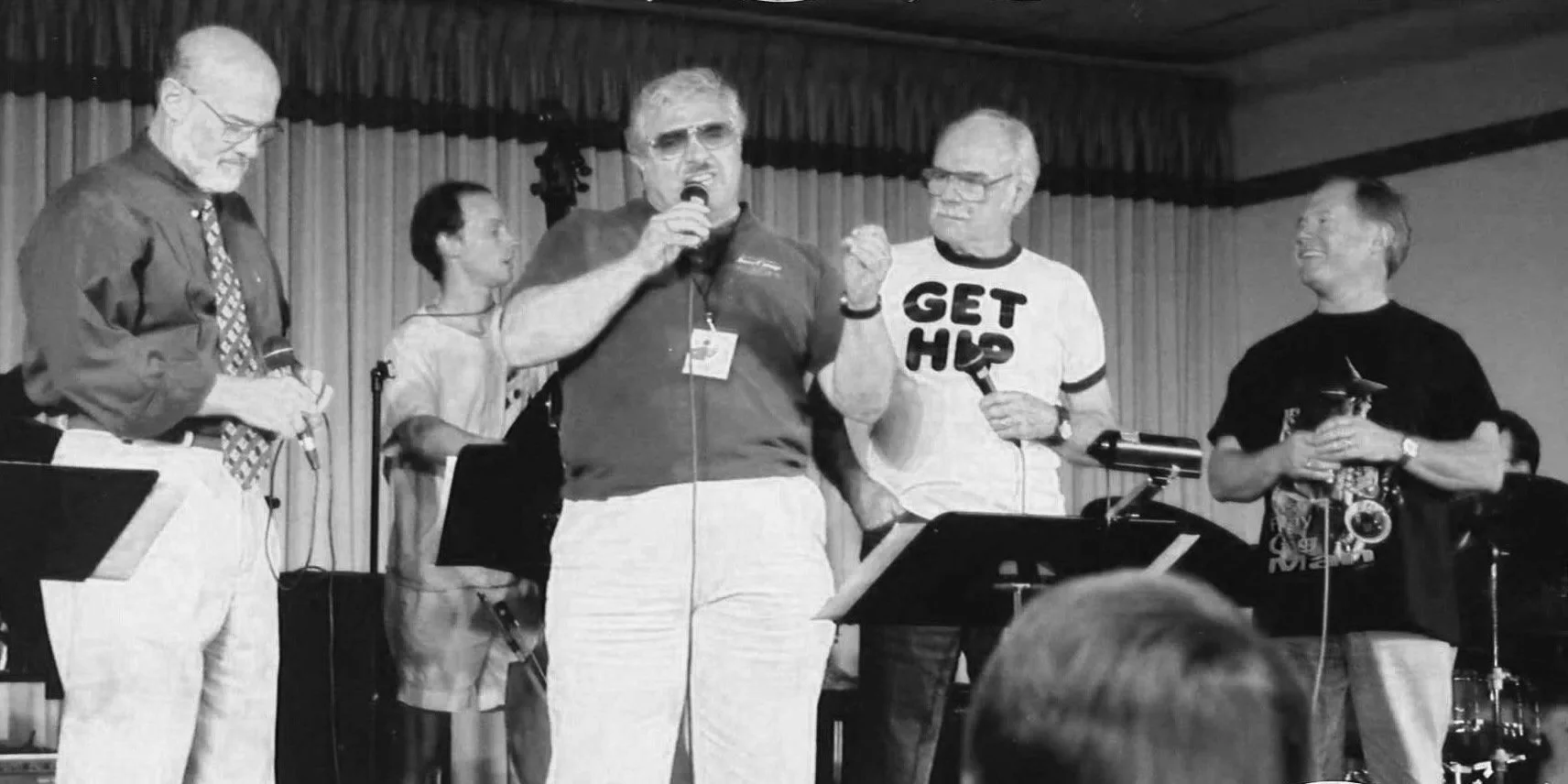 Group of people on stage, one man speaking into a microphone and wearing a shirt that says 'GET HIP', others holding microphones or musical instruments, with a curtain background.