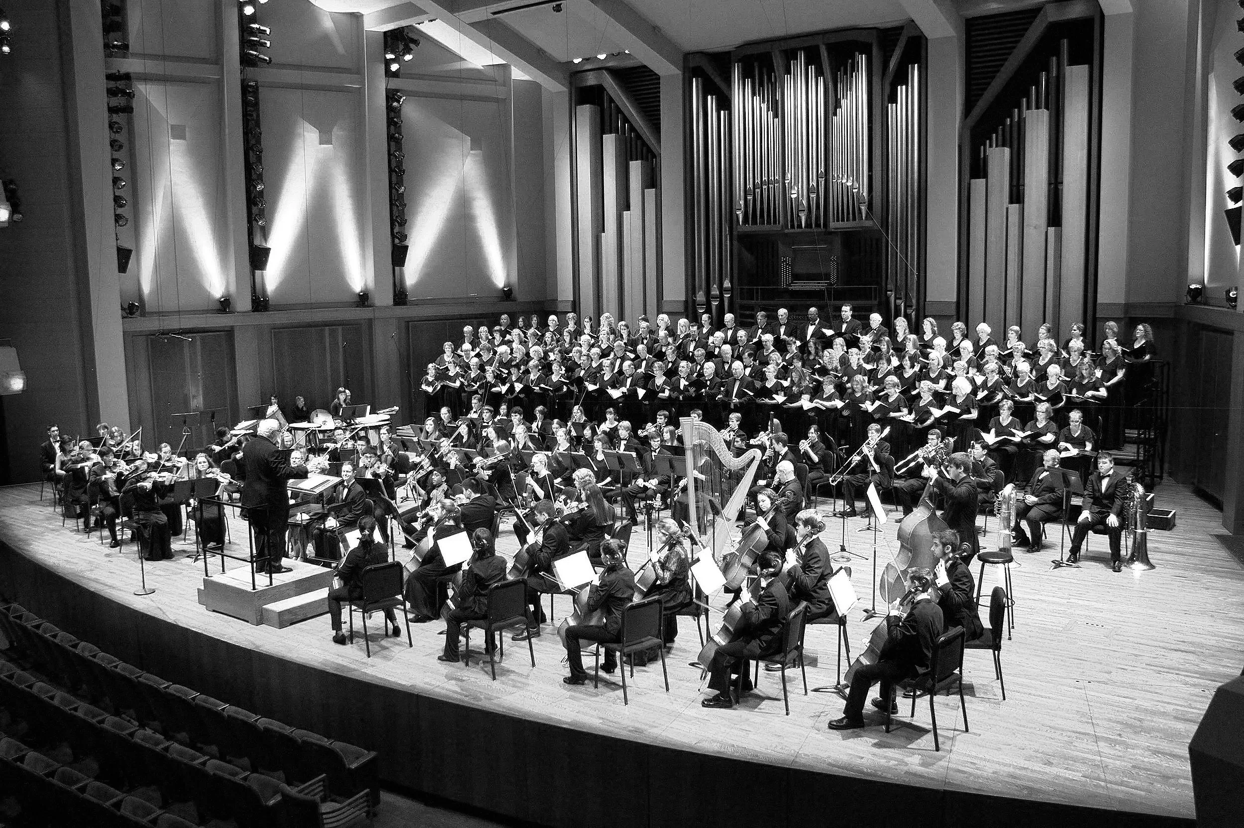 An orchestra performs on stage with musicians playing various instruments, directed by a conductor. A choir stands behind the orchestra in a concert hall.