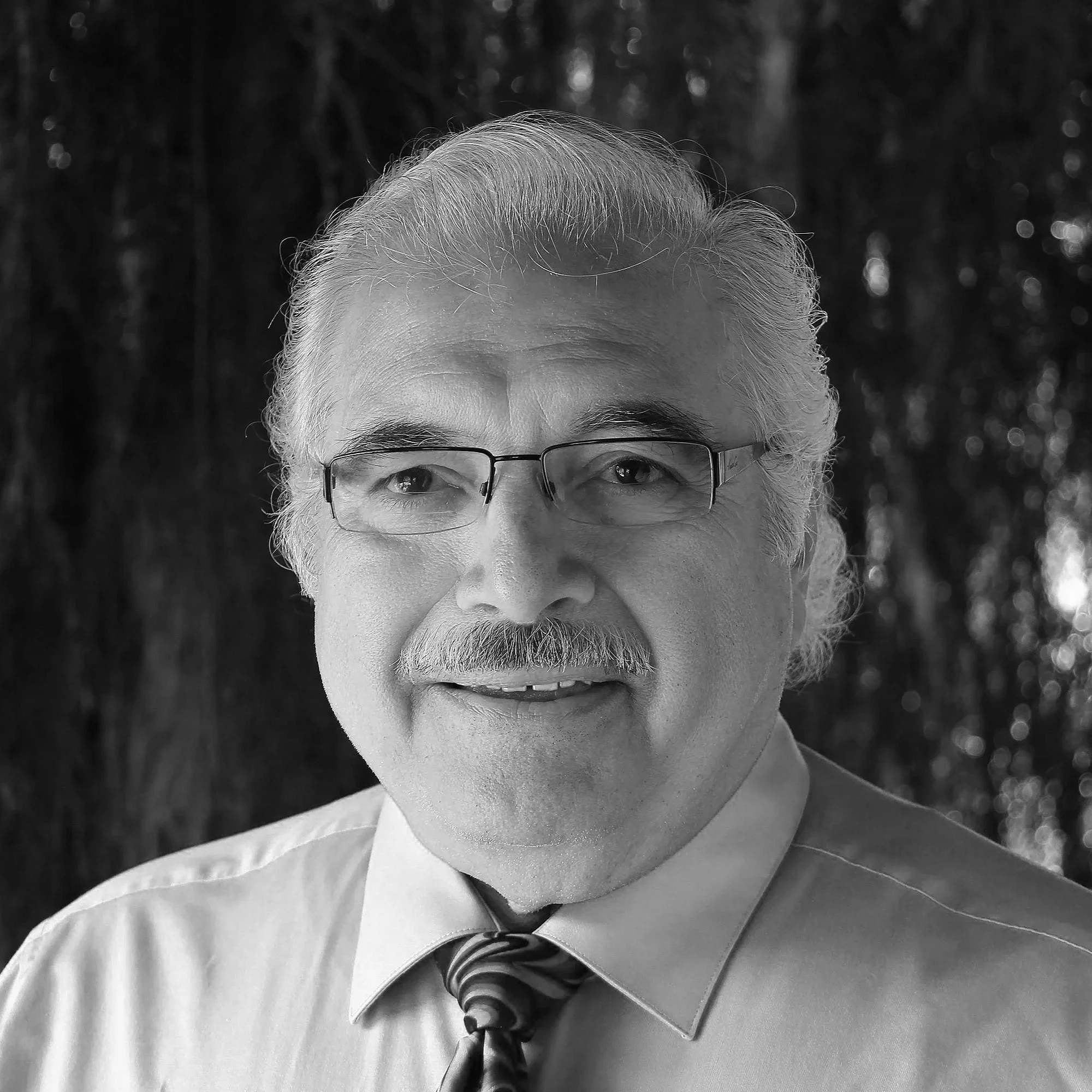 Black and white portrait of an older man with glasses, a mustache, and gray hair, wearing a collared shirt and a patterned tie, outdoors with blurred trees in the background.