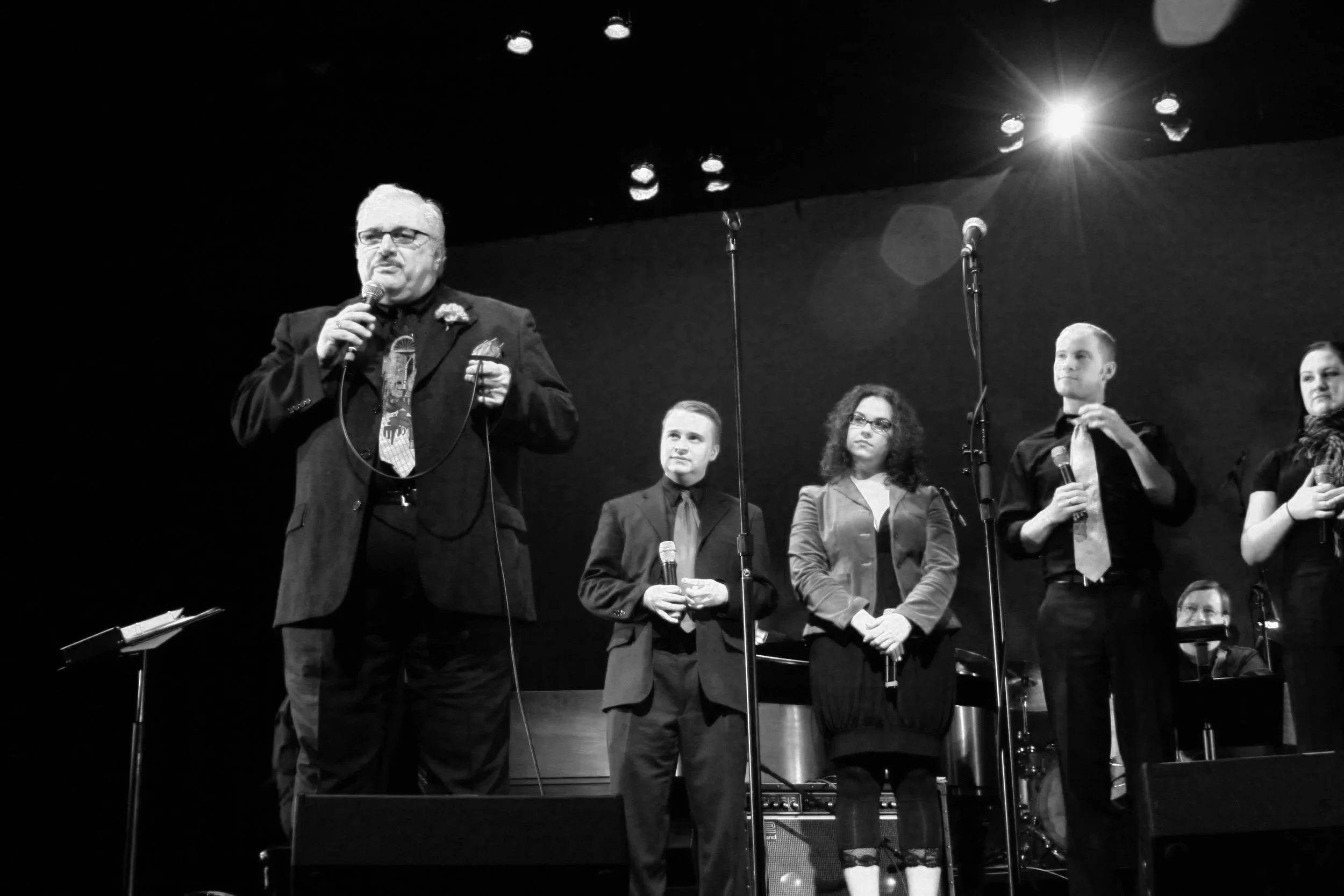 A man with glasses and a mustache holding a microphone speaks on stage, with four young people standing behind him, also holding microphones, in a black-and-white setting with stage lights and musical instruments.