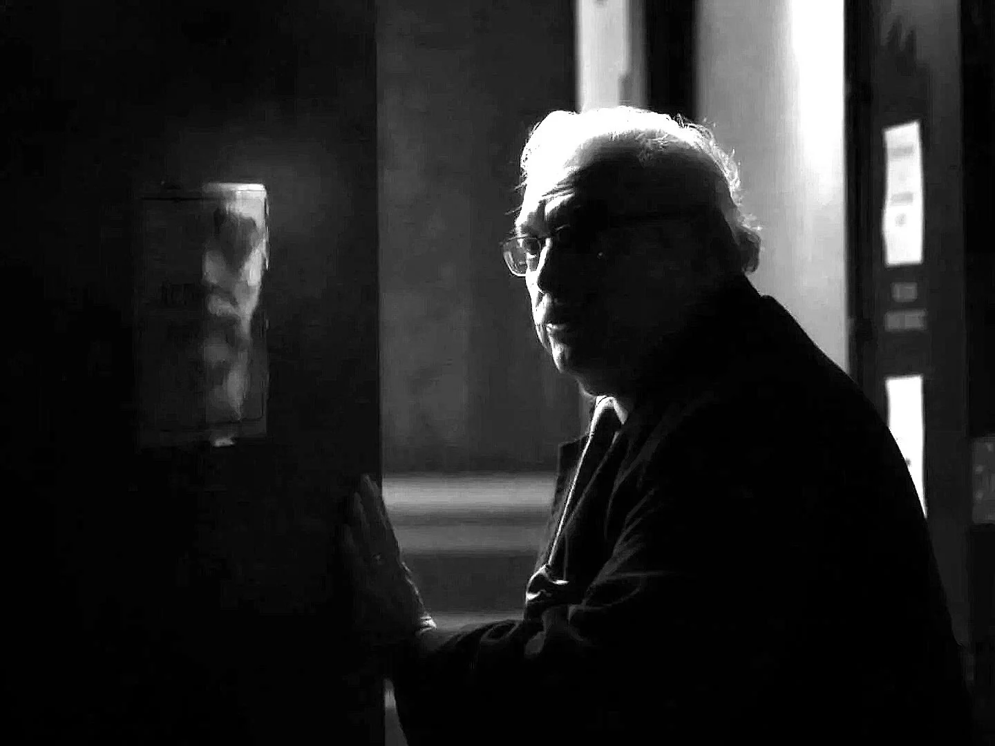 Black and white photo of an elderly man with glasses, sitting in front of vending machines in a dimly lit setting.