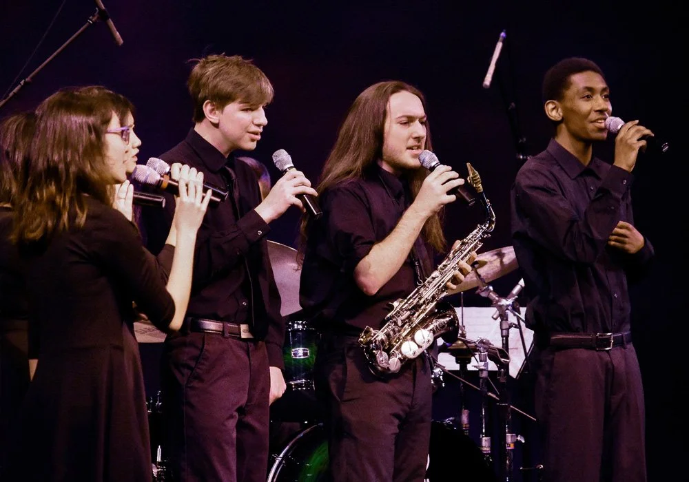 Four young musicians performing on stage, each holding a microphone, with a drum set and other musical equipment in the background.