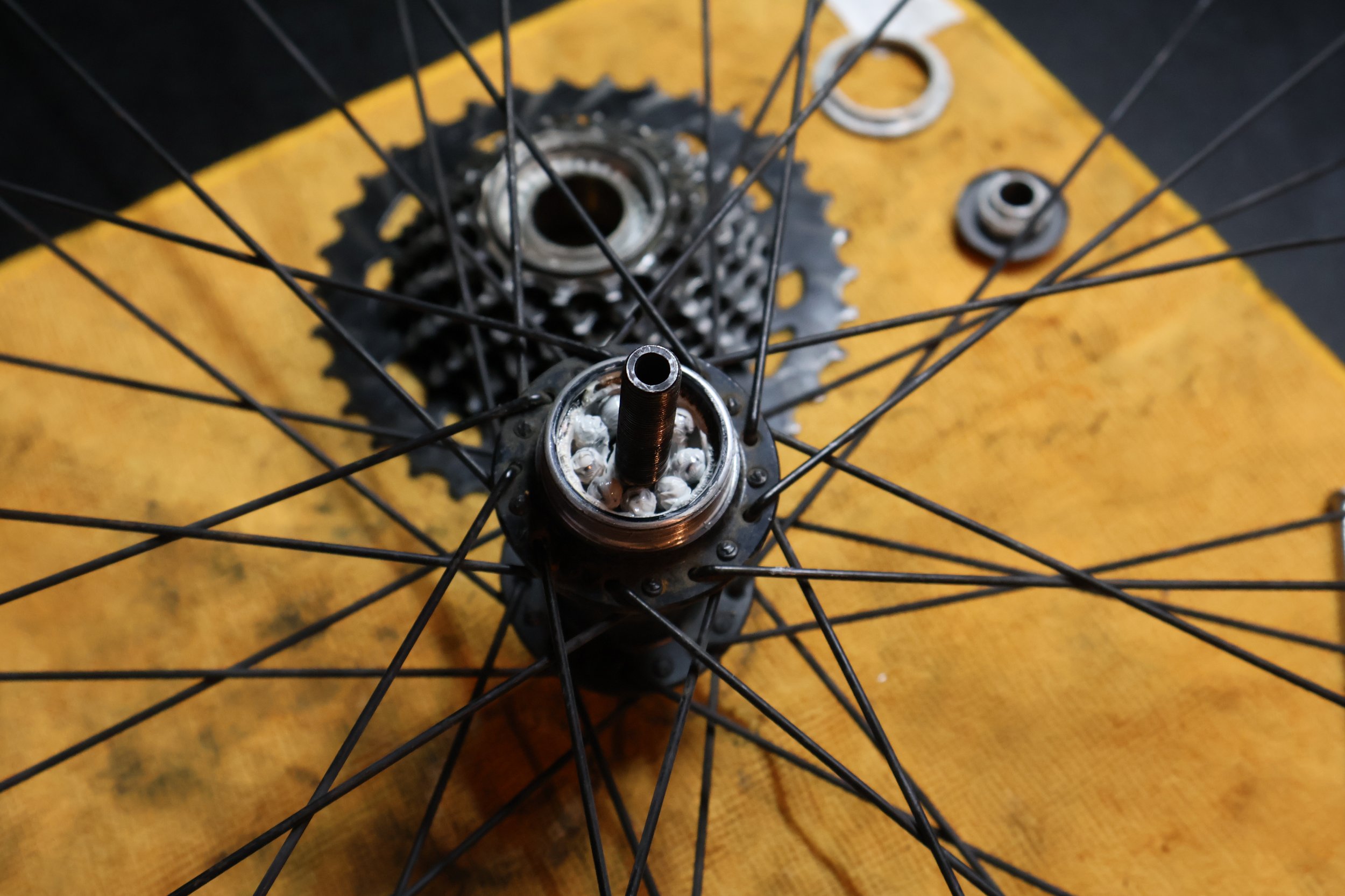 A close-up of a bicycle wheel hub with spokes, part of a bike wheel, and a cassette gear, placed on a yellow wooden surface.