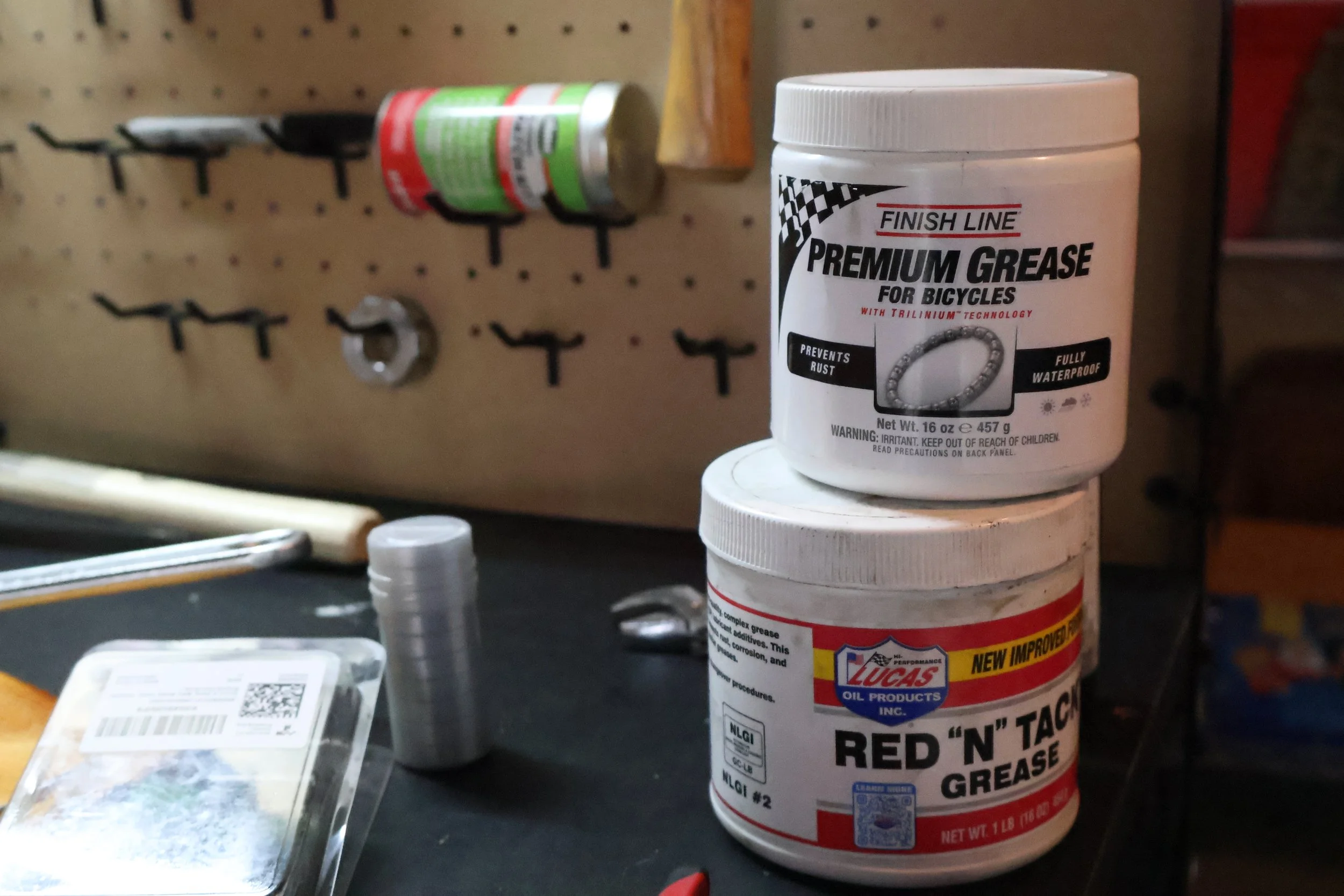 Containers of bicycle greases, one labeled 'Finish Line Premium Grease for Bicycles' and the other labeled 'Lucas Oil Products Red N Tag Grease,' on a black workbench in a workshop. The background has a pegboard with tools.