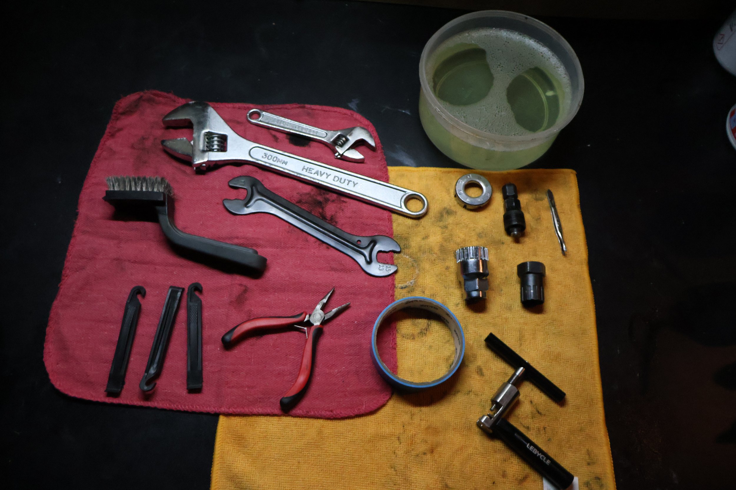 Assorted bicycle repair tools and parts on a red and yellow cloth with a cup of green liquid nearby.