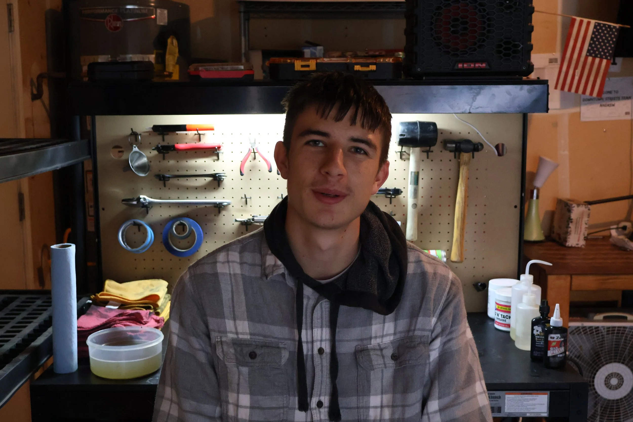A young man with short dark hair and wearing a plaid shirt sits in a workshop with tools and supplies on a pegboard and workbenches behind him.