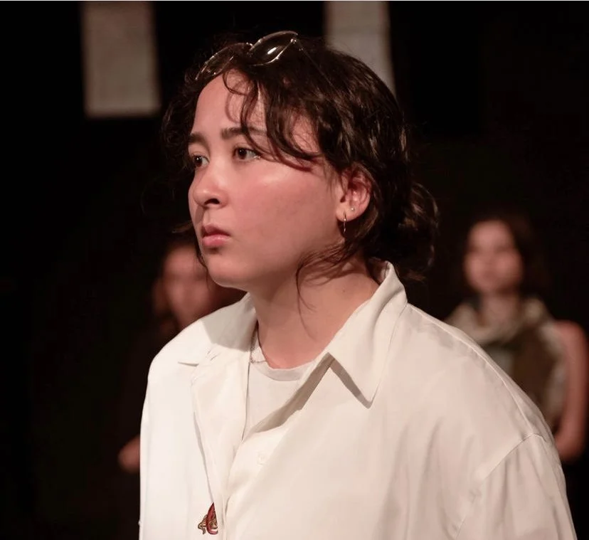 A young person with wavy brown hair, wearing a white shirt, standing in a dimly lit room, looking serious or contemplative.