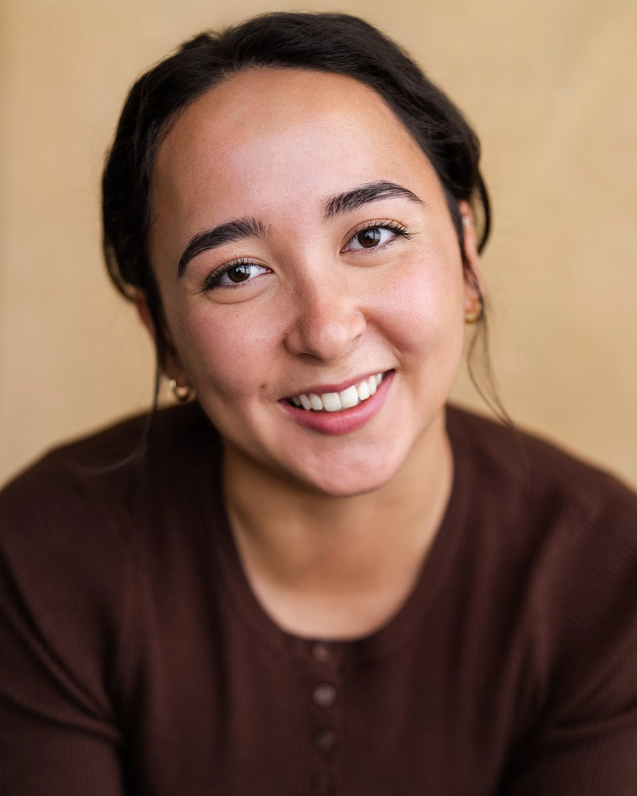 Close-up of a young woman with dark hair, smiling, wearing a brown top, against a beige background.