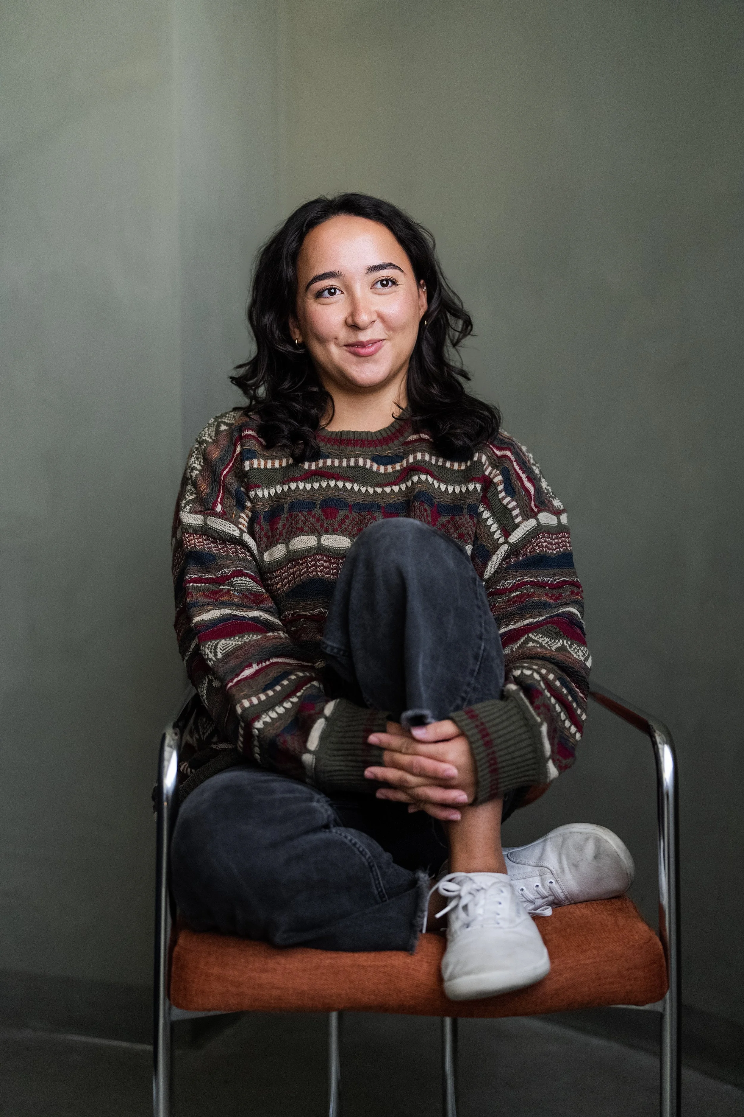 A young woman with dark wavy hair sitting on an orange cushioned chair against a plain green wall, wearing a patterned sweater, black jeans, and white sneakers, with her legs crossed and hands resting on her knee.