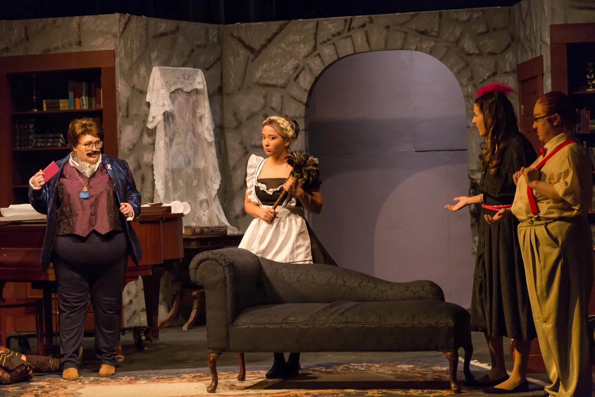 The image shows a theatrical play scene with four actors in costume on a set that resembles a vintage living room with stone walls, a sofa, bookshelves, and a large arched door in the background.