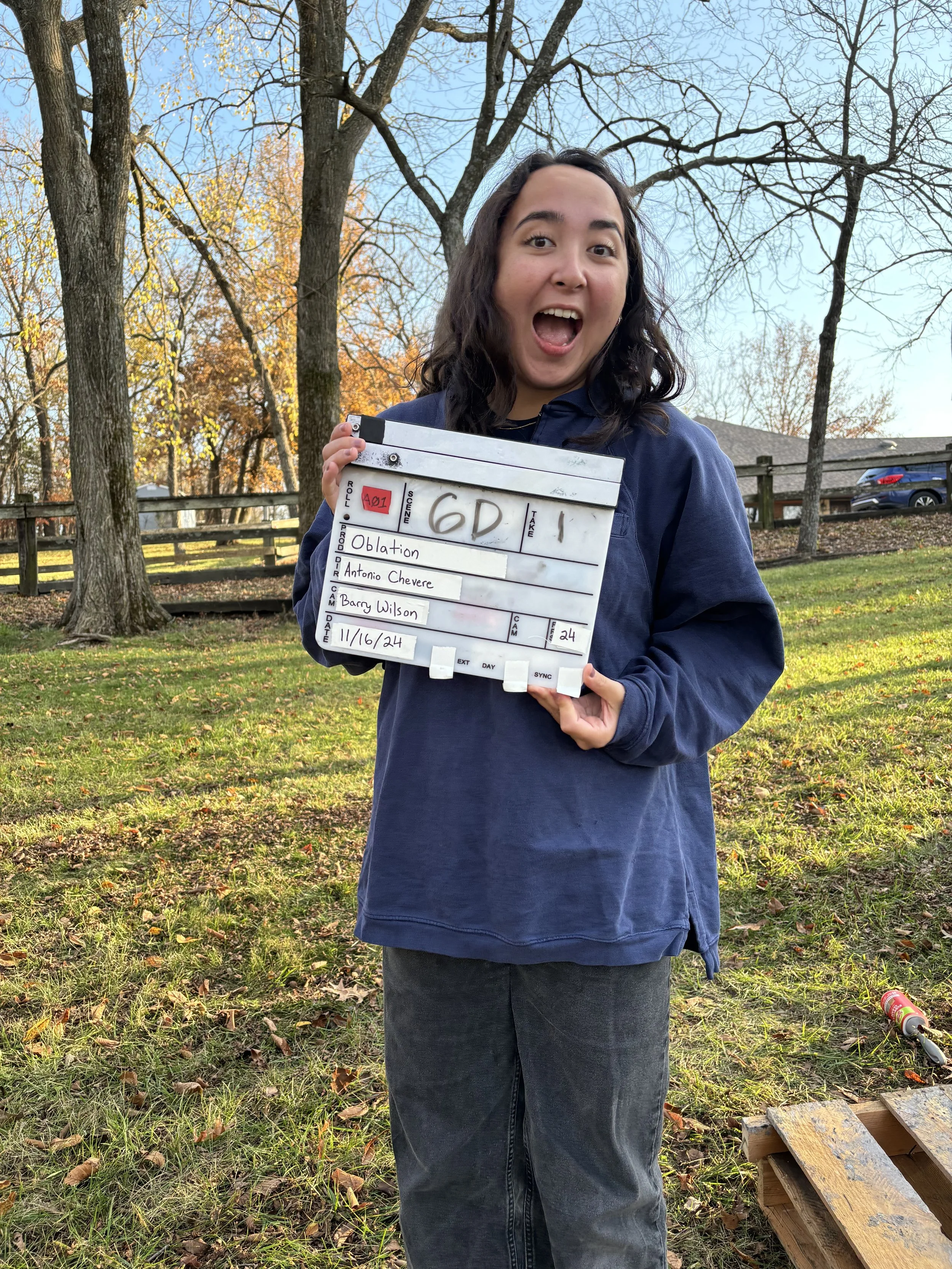 A young woman with dark hair, wearing a navy jacket, standing outdoors in a park with trees and fallen leaves, holding a clapperboard with writing on it and smiling with her mouth open.