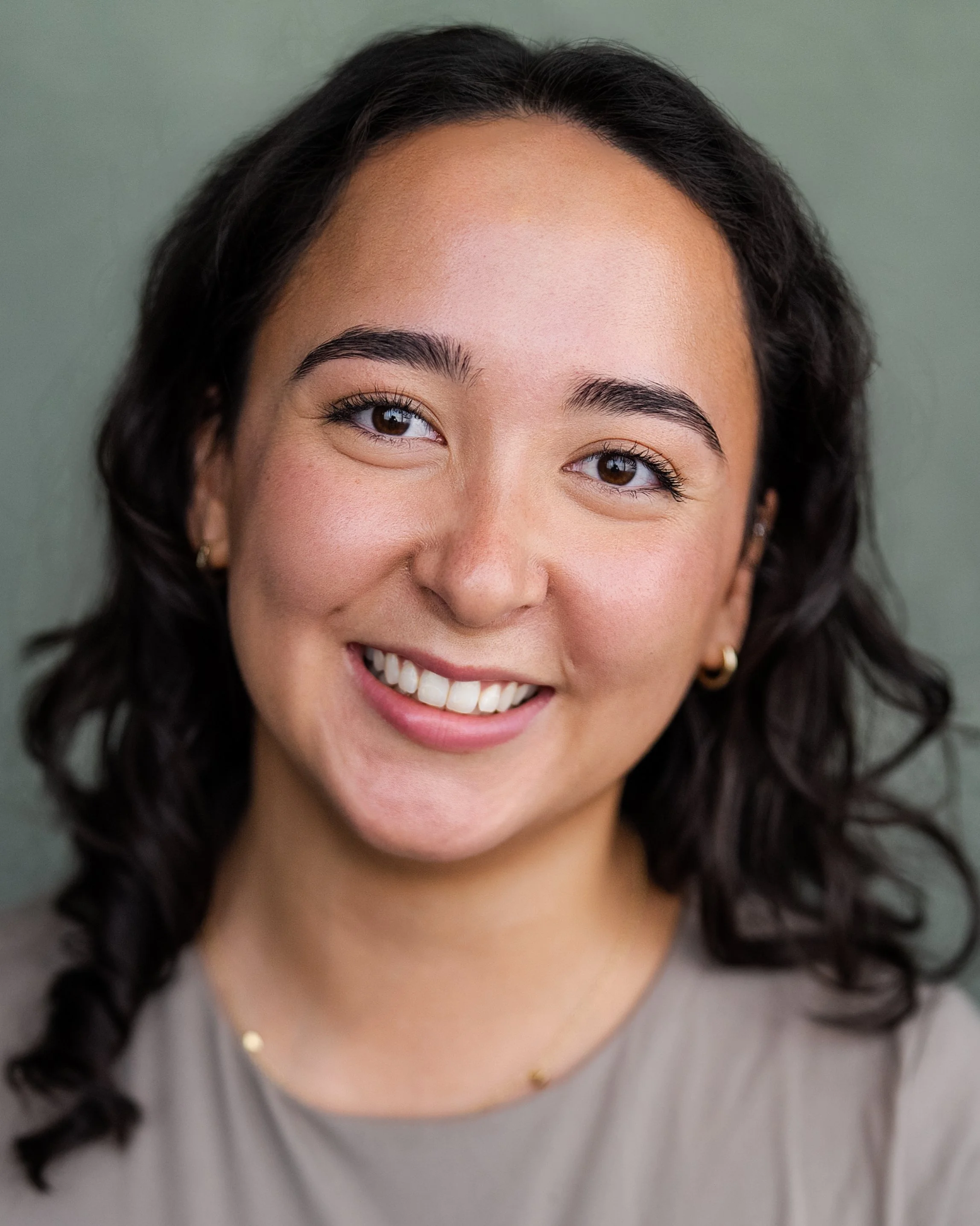 Close-up portrait of a smiling young woman with dark curly hair, wearing gold earrings and a light-colored top, against a muted green background.