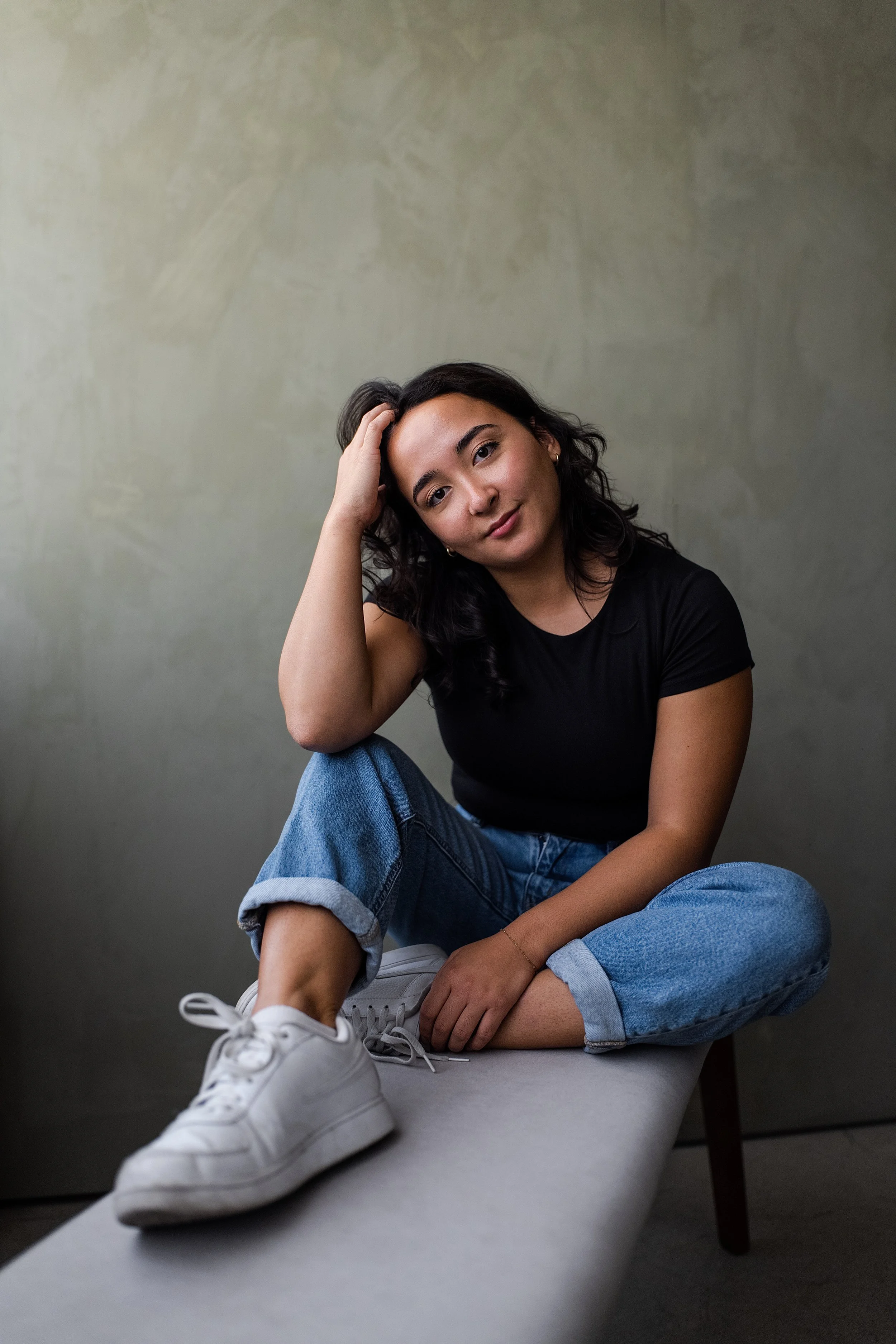 A young woman with dark hair, wearing a black t-shirt, blue jeans, and white sneakers, sitting on a bench with a neutral background, resting her head on her hand and looking at the camera.