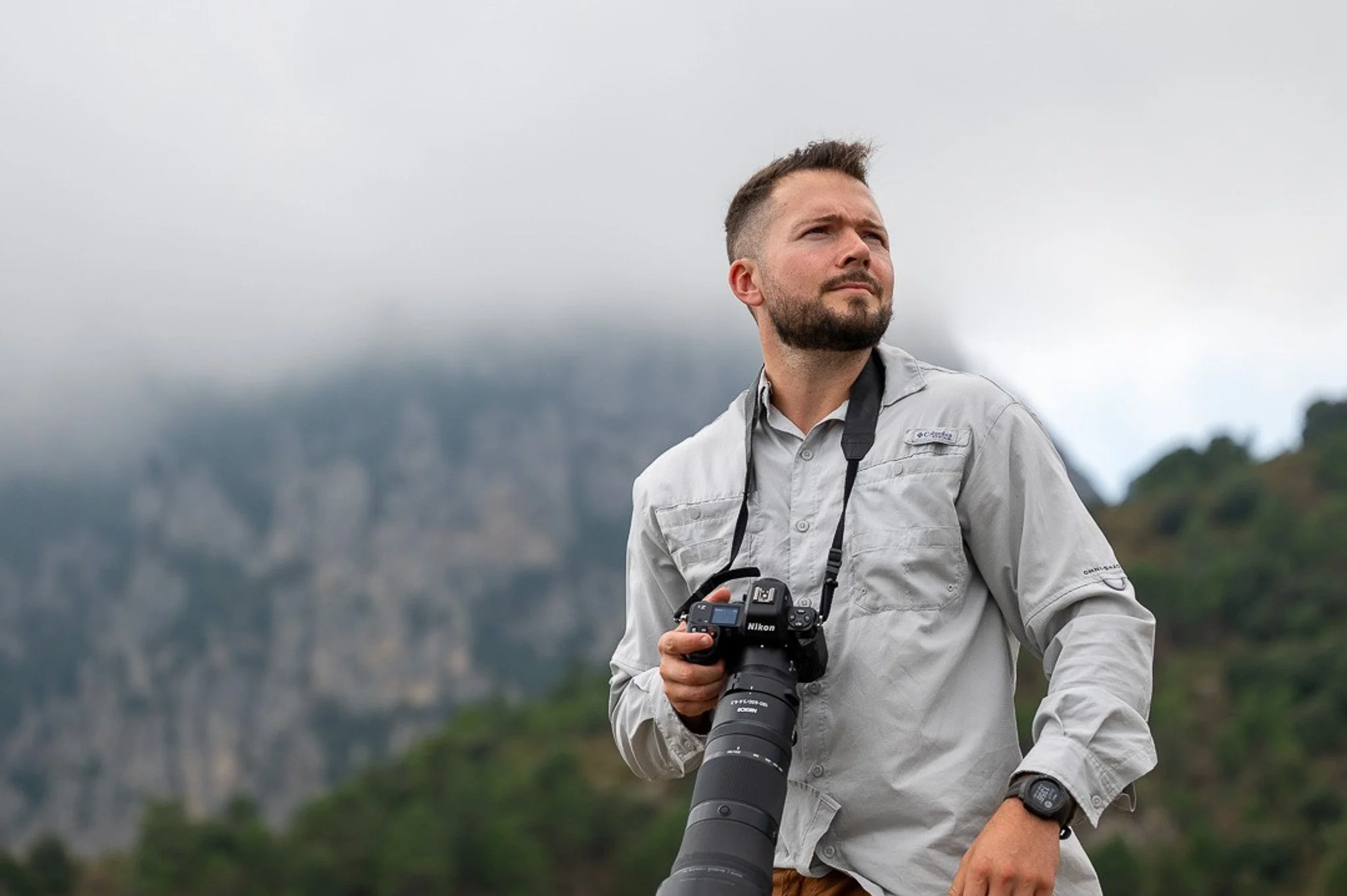 Travis Layton known as Trav in the Bush standing in front of a mountain holding his Nikon camera.
