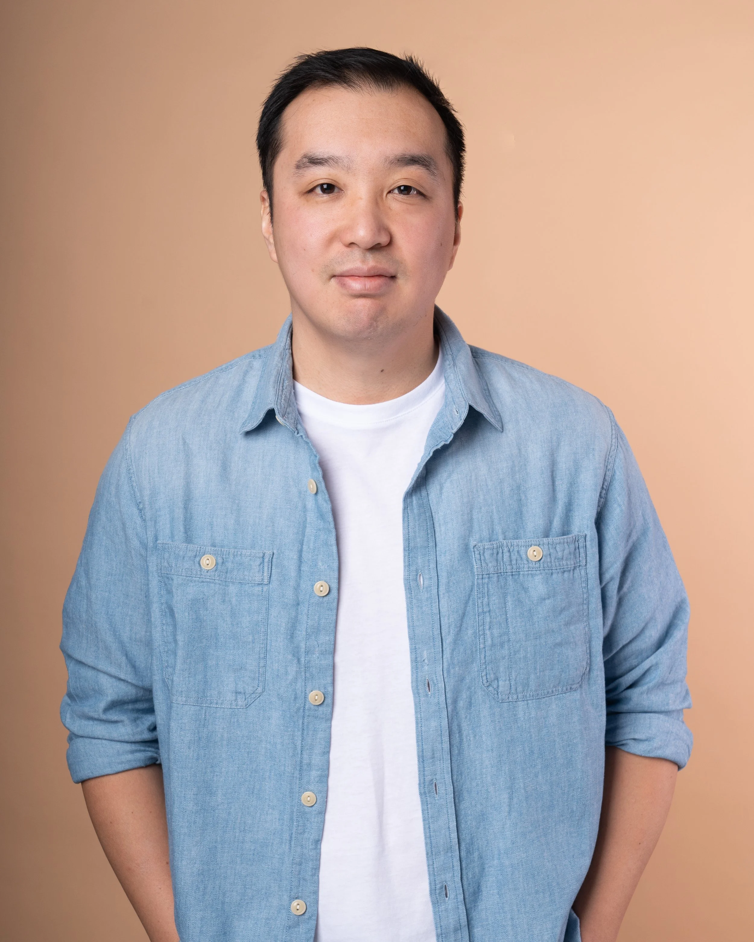 A person wearing a light blue denim shirt over a white t-shirt, standing against a beige background.