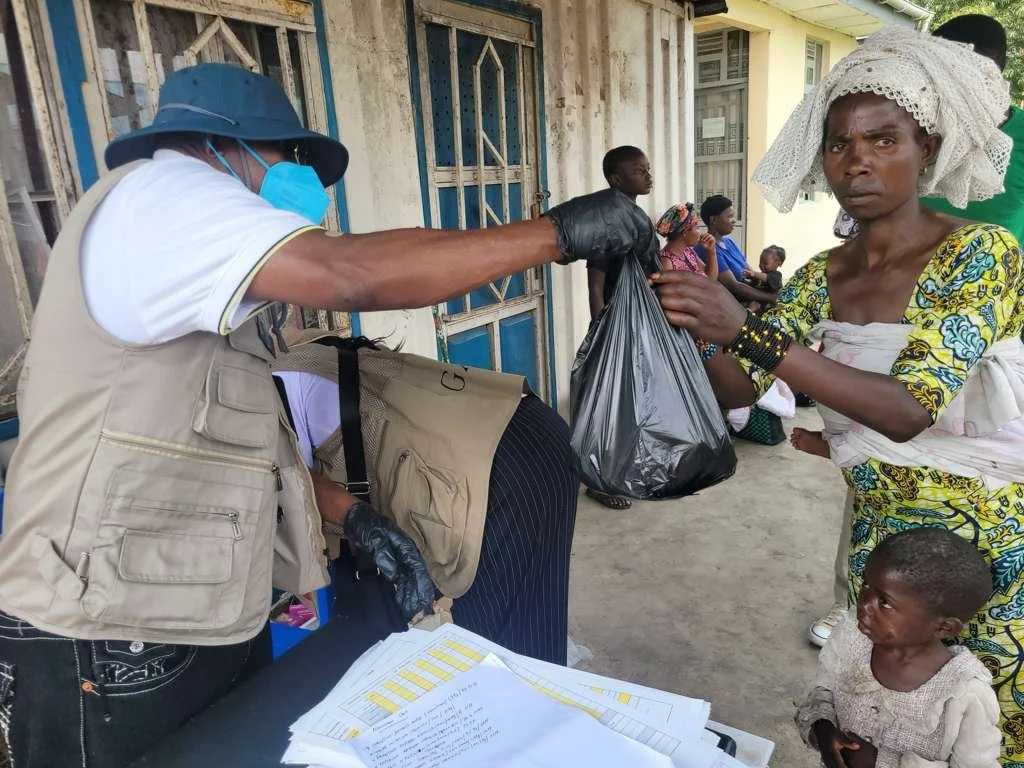A woman in traditional clothing receiving a black plastic bag from a man in a face mask and gloves, outside in a community setting with other people sitting or standing in background.