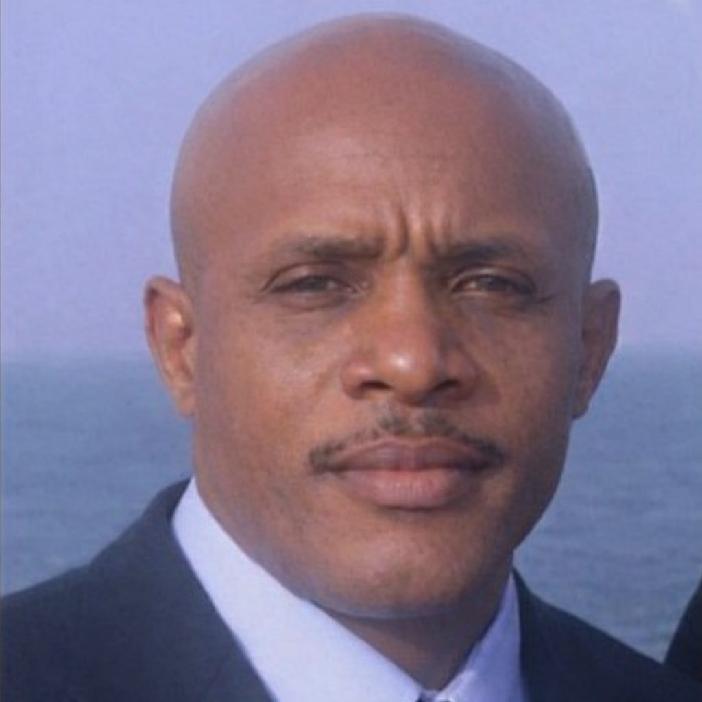 Close-up photo of a bald man with a serious expression, wearing a dark suit and white shirt, with a body of water in the background.