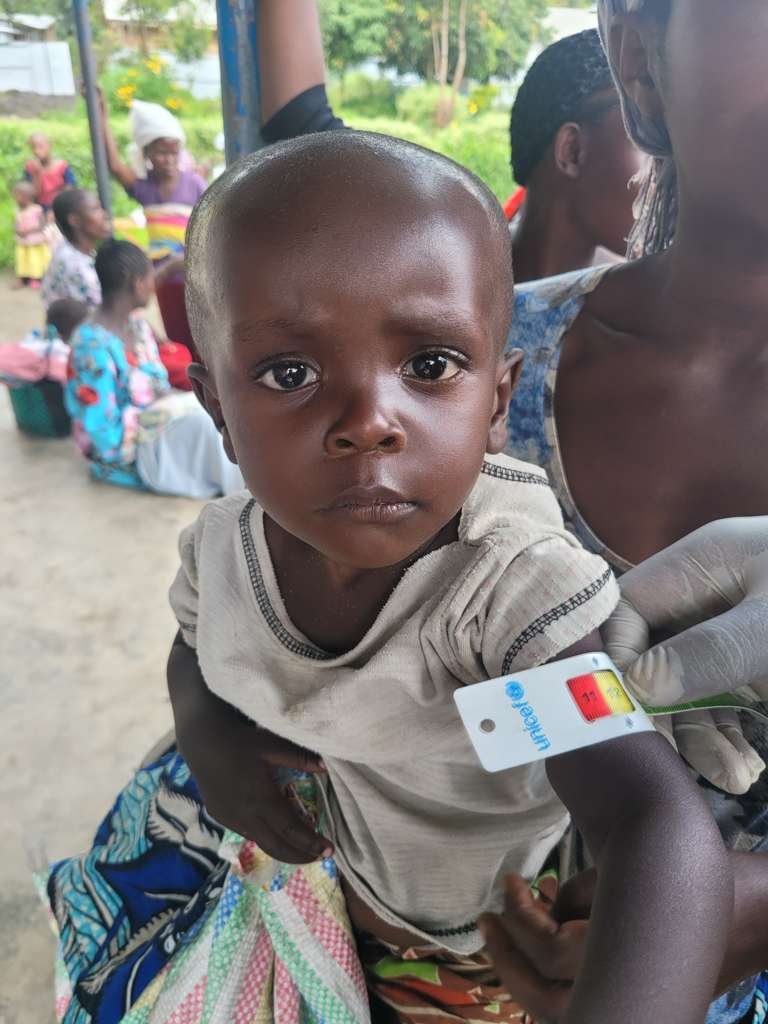 A young child receives a vaccination from a healthcare worker in an outdoor setting. Several other children and adults are visible in the background.
