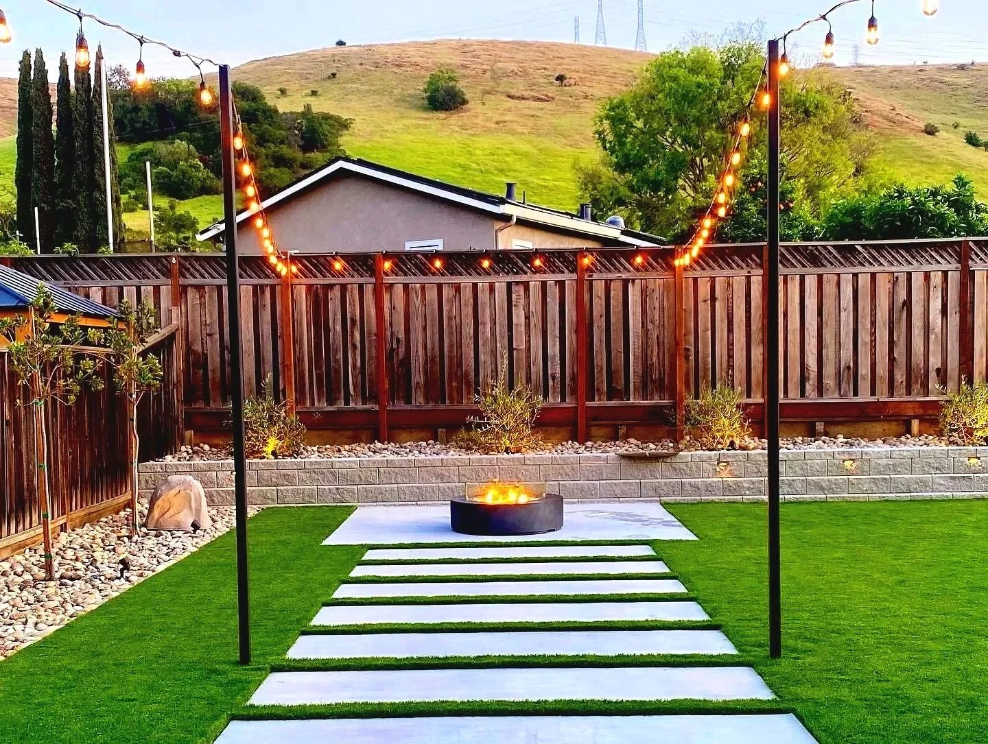 Backyard with artificial grass, stepping stones, a fire pit, string lights, wooden fence, planted bushes, and hillside in the background.