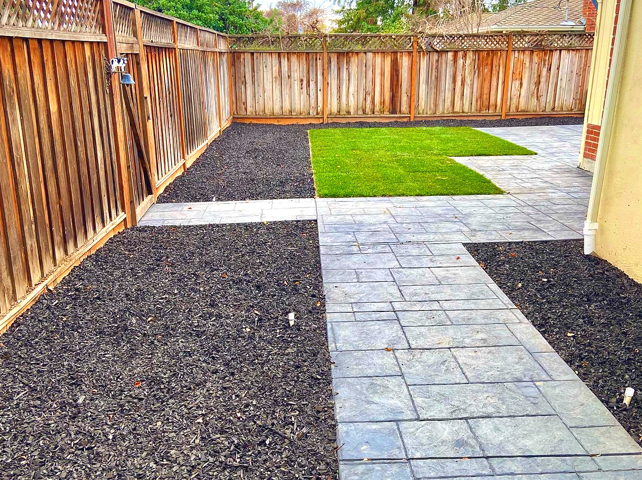 A backyard with a wooden fence, stamped concrete patio, areas of gravel, and a section of green grass.