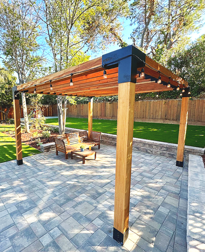 Wooden outdoor pergola with string lights and wooden benches on a paver patio, overlooking a backyard with a grassy lawn and trees.