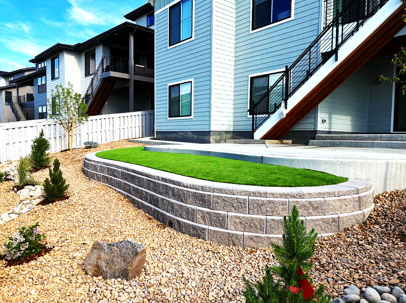 Exterior view of a modern multi-story residential building with a landscaped backyard featuring turf on a stone retaining wall, gravel ground cover, small shrubs, and young trees, with a wooden fence in the background.
