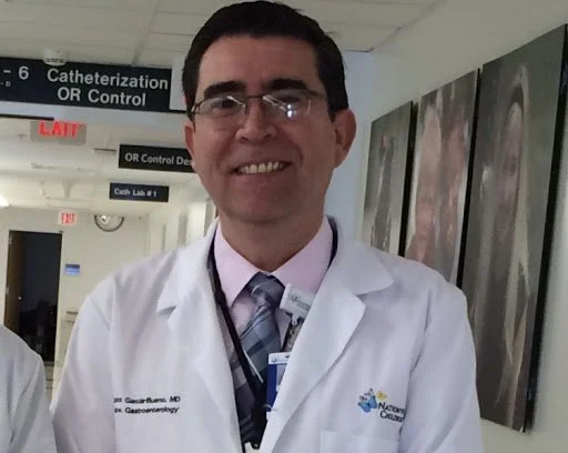 Un médico sonriente con bata blanca en un hospital, posiblemente en un pasillo de la sala de radiología o radioterapia.