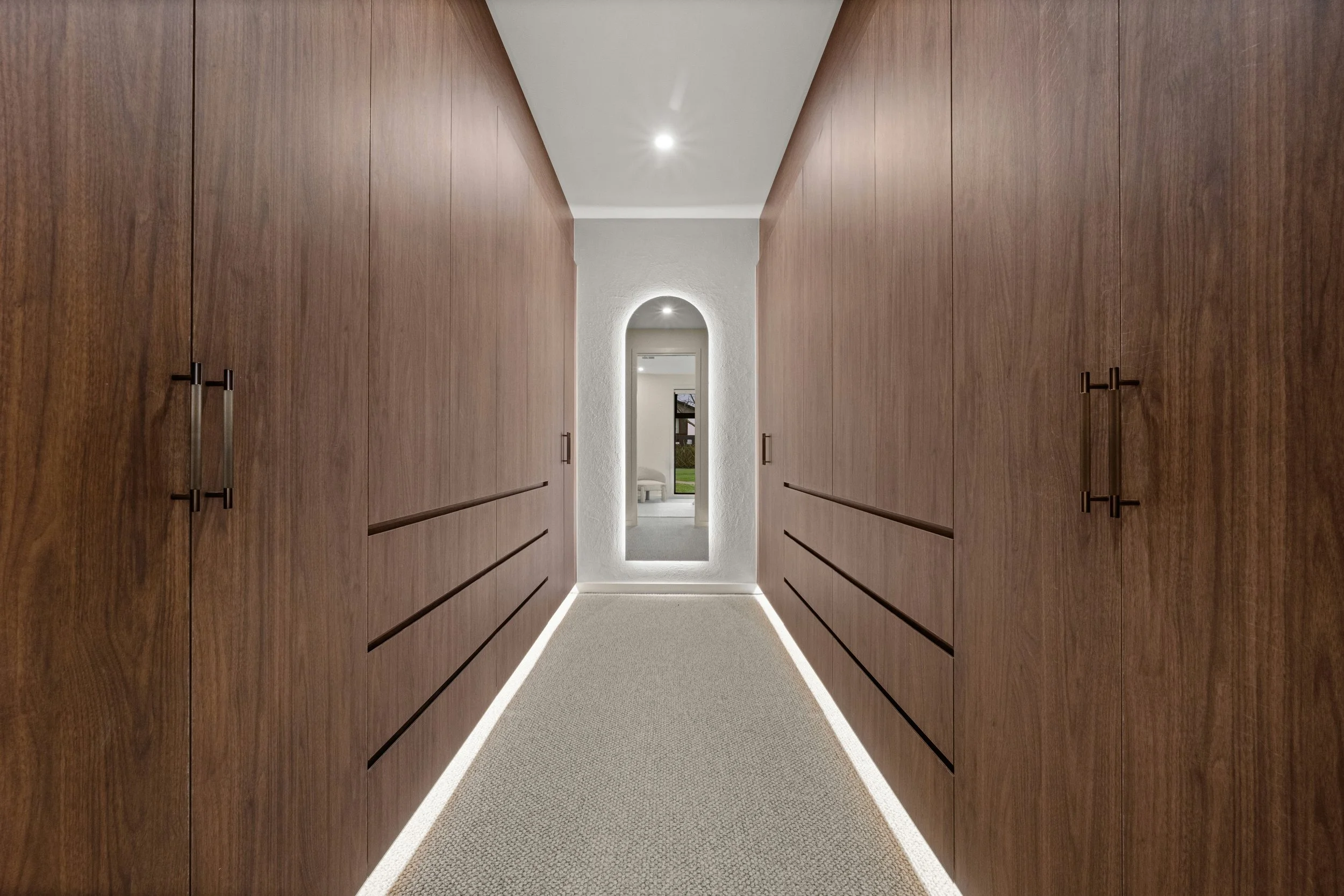 Modern walk in robe with wooden cabinetry on both sides, arched mirror at the end, and soft lighting along the floor.