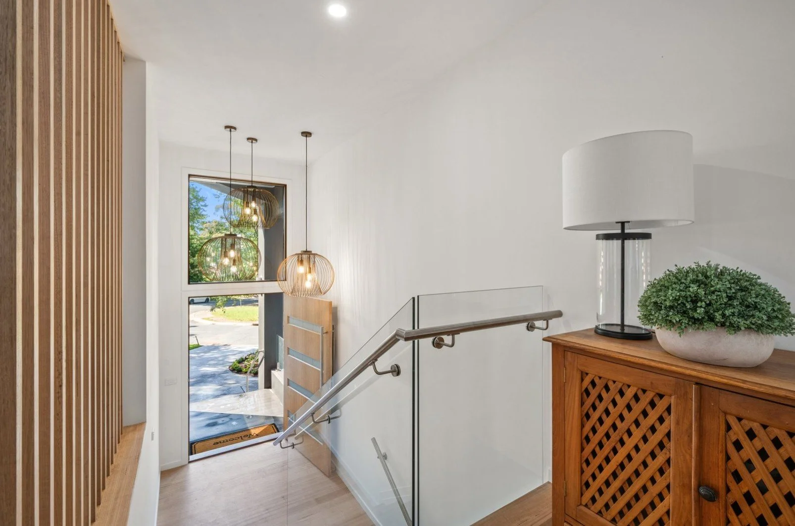 Interior view of a modern entryway with a staircase, wooden sideboard with a lamp and potted plant, and hanging pendant lights near a window with trees outside.