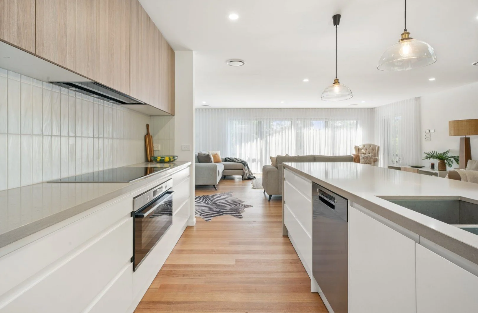 Open-concept kitchen and living room with white cabinets, light wood floors, and large windows with sheer curtains, furnished with sofas, a zebra print rug, and decorative plants.