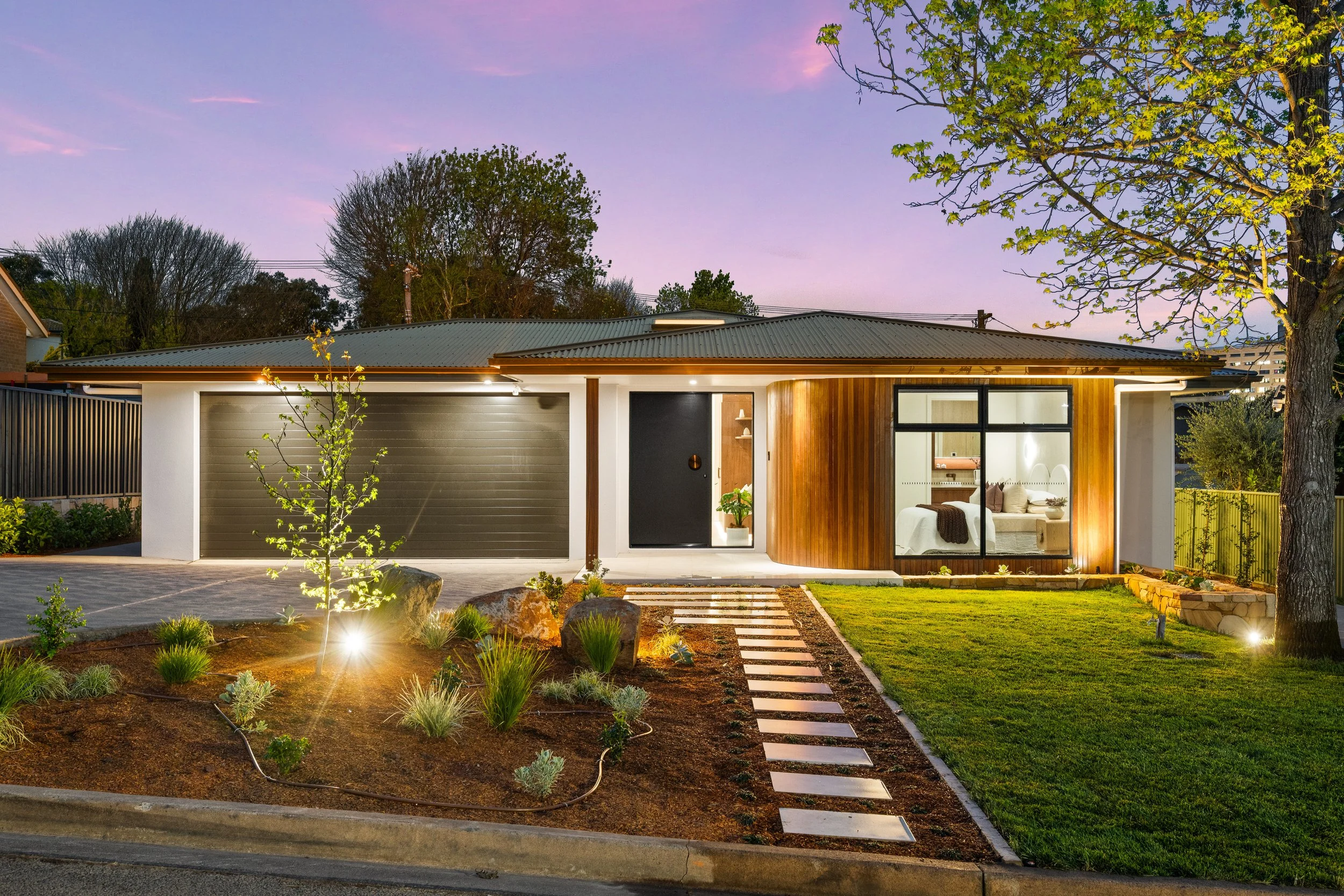 Modern house with a gray garage door and a wooden accent wall, large windows revealing a cozy interior, surrounded by a landscaped yard with small plants, rocks, and pathway lights, under a twilight sky with trees in the background.