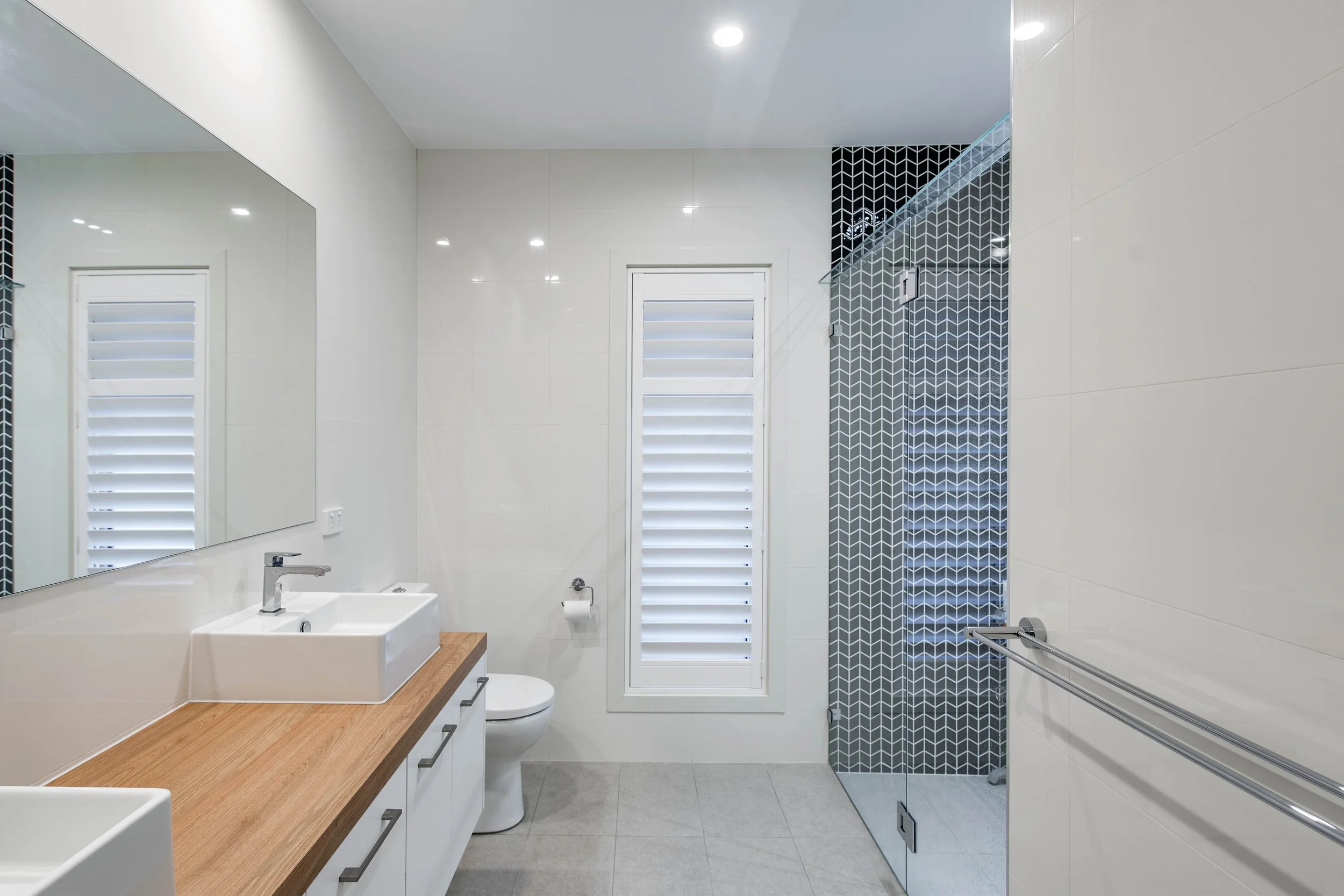 Modern bathroom with white walls, a mirror, a white sink with a wooden countertop, a toilet, a window with white slats, and a glass-enclosed shower with black and white geometric tiles.