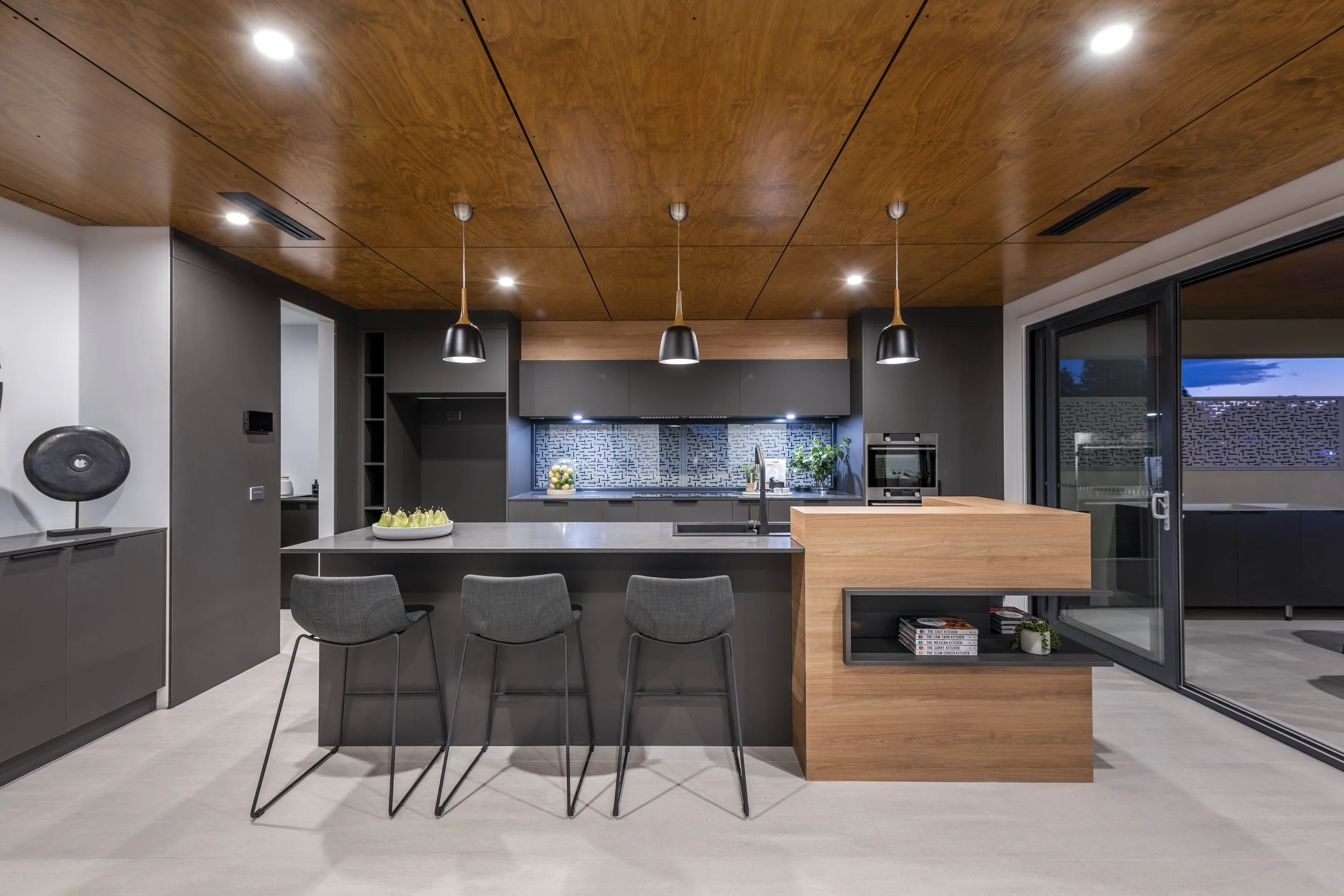 Modern kitchen with wooden ceiling, grey cabinets, black pendant lights, white countertops, and a sliding glass door. There are four grey chairs at the island, with a bowl of fruit on top. The backsplash features a geometric pattern, and there's a pl