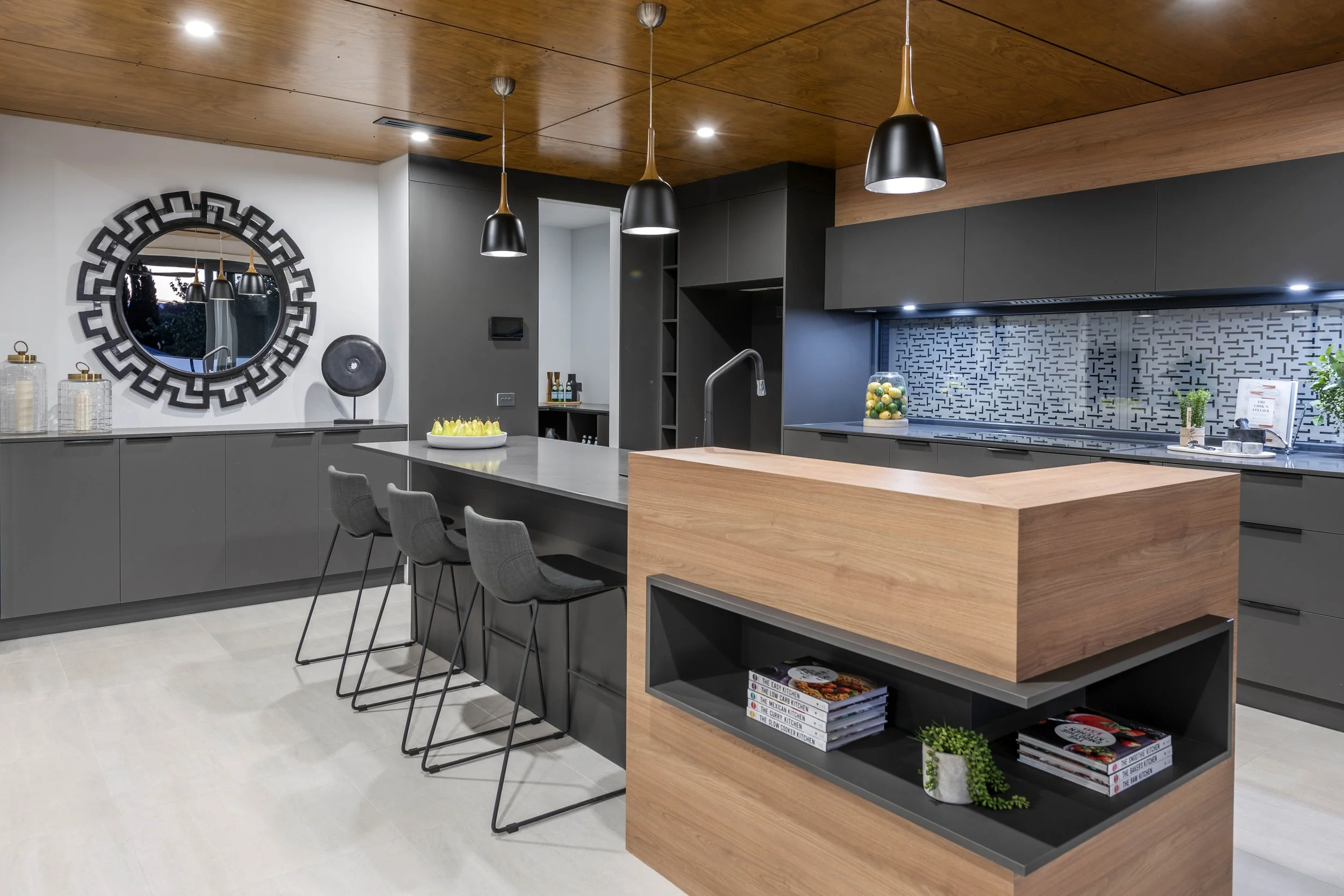 Modern kitchen with gray cabinets, a wooden ceiling, and black pendant lights. There is a large circular mirror on the wall, a kitchen island with a gray countertop, and shelves stocked with cookbooks and decor. Plants and decorative jars are also vi