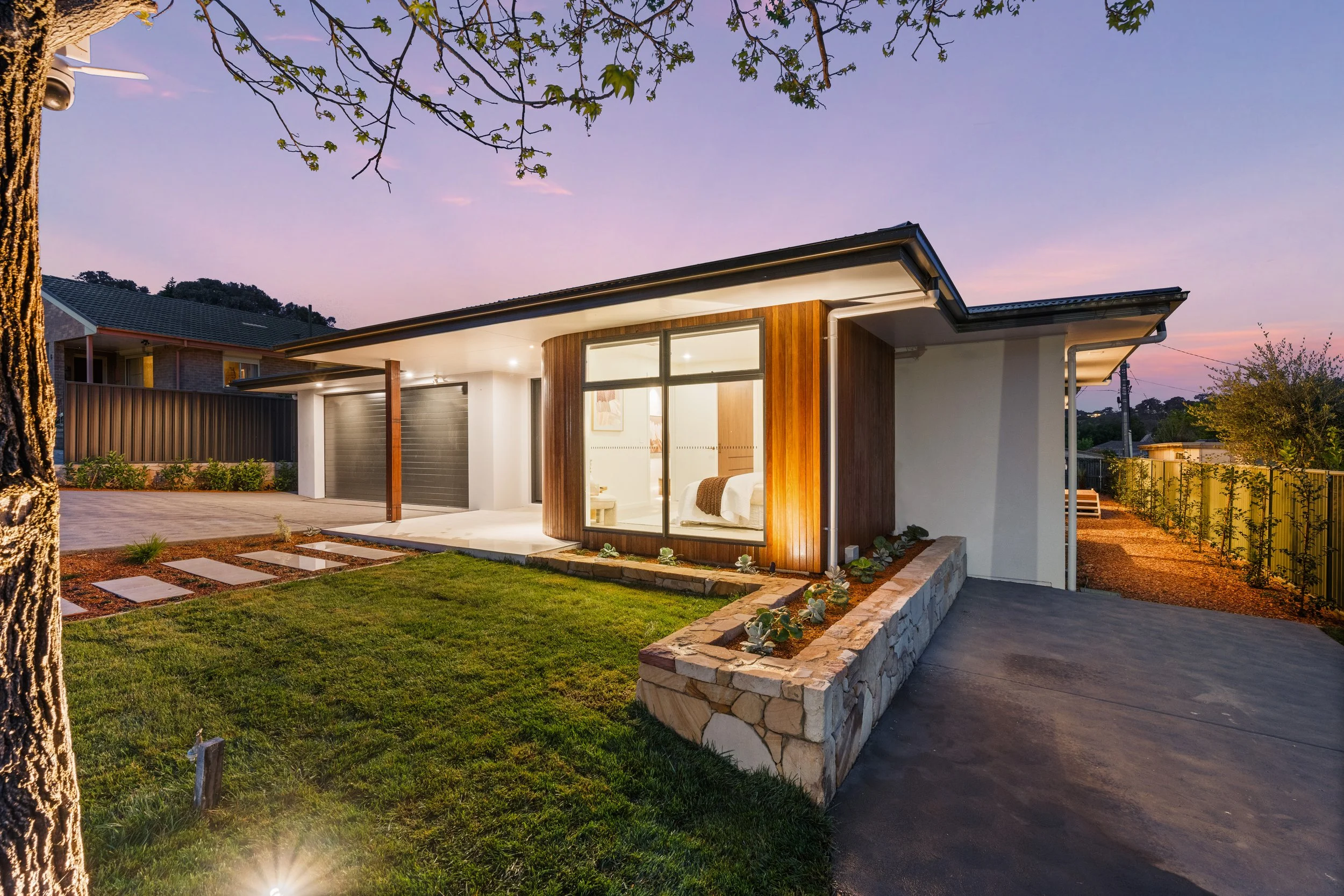 Modern house with large glass windows and a mix of white and wooden exterior, illuminated by outdoor lighting at dusk, with a small lawn, stone pathway, and a tree in the foreground.