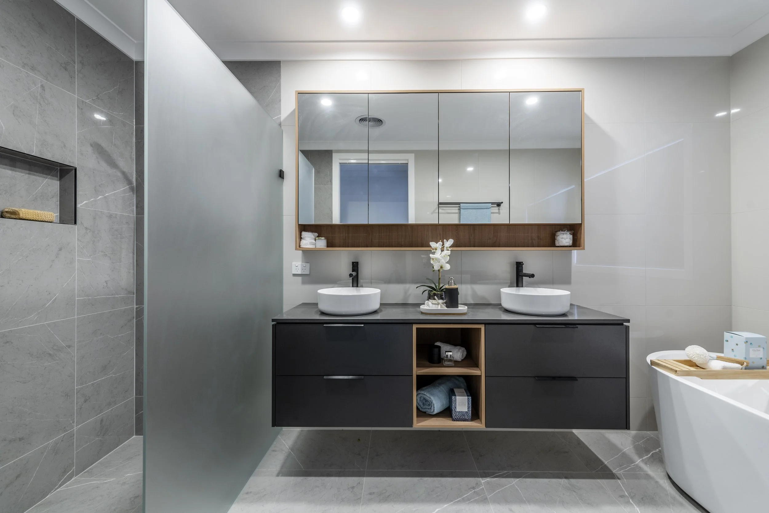 Modern bathroom with grey marble tiles, a large mirror above a double sink vanity, and various toiletries and decor items on shelves and counters.