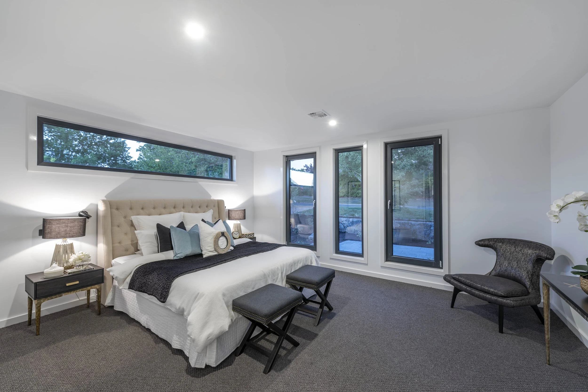 Modern bedroom with a large bed, beige tufted headboard, white bedding, black throw blanket, decorative pillows, two black nightstands with lamps, and floor-to-ceiling windows showing trees outside.