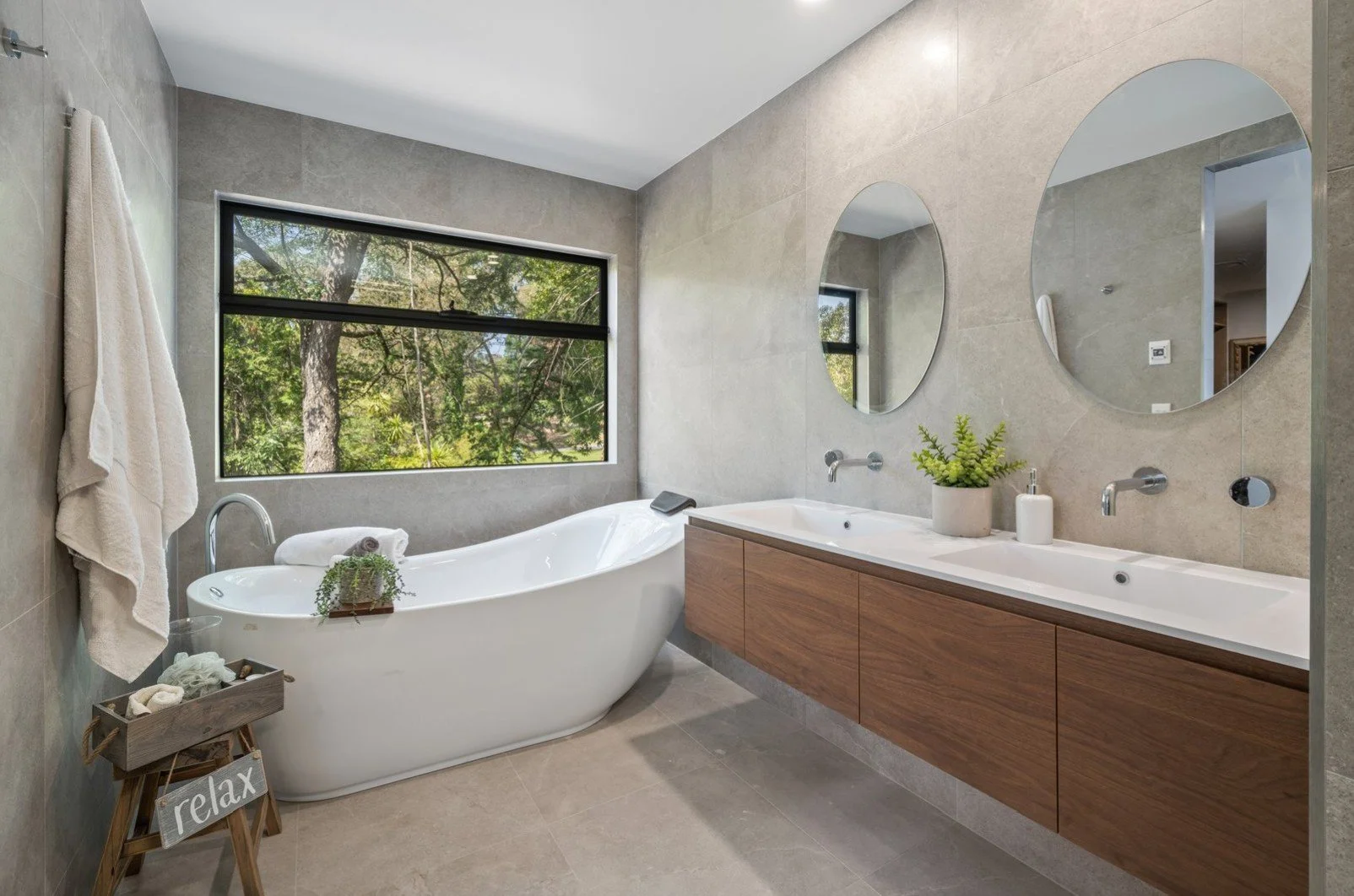 Bathroom with a white bathtub by a large window, two round mirrors above a wooden vanity with a white sink, potted plant, soap dispenser, towels, and decorative items.