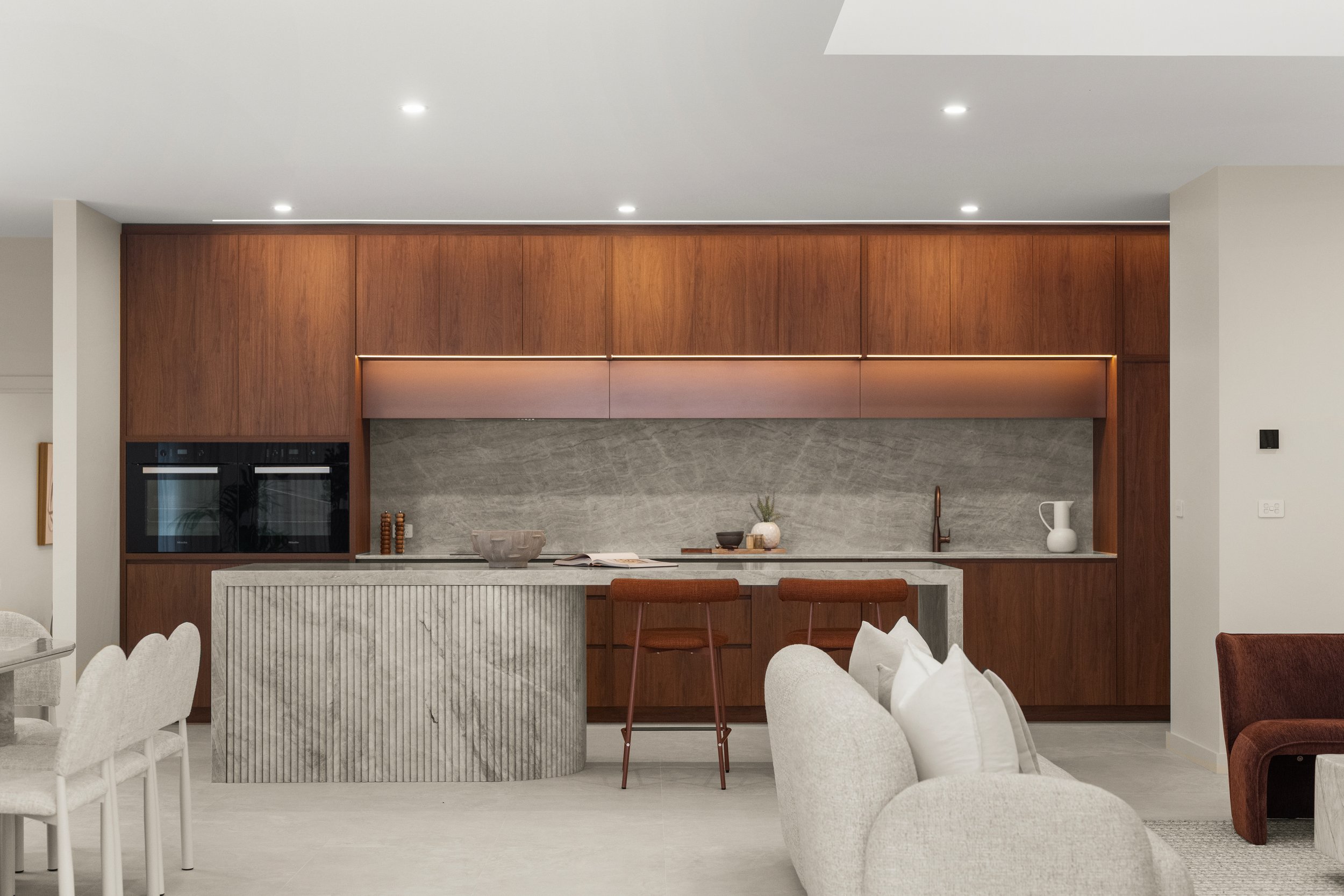 Modern kitchen with wooden cabinetry, grey marble backsplash, and a kitchen island with striped vertical design. White dining chairs are visible in the foreground, and decorative items like bowls, vases, and plants are on the countertop.