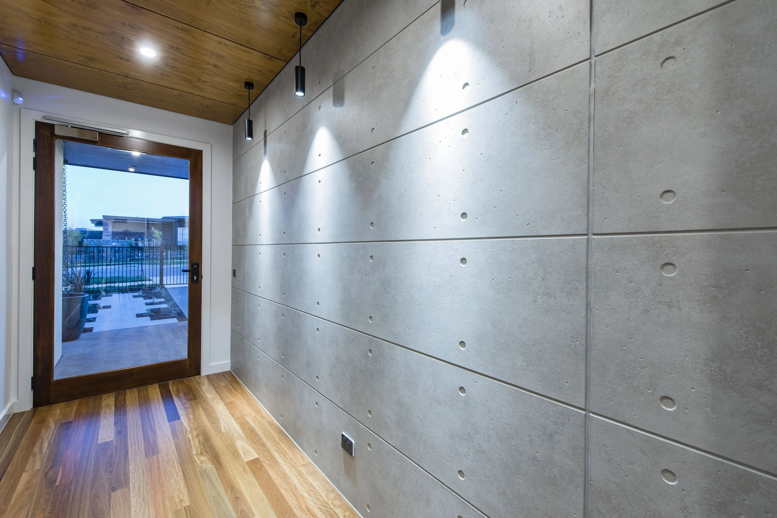 Interior of a modern room featuring a concrete wall with round holes and a wooden ceiling, a wooden floor, and a door with glass leading outside with plants and a building visible.